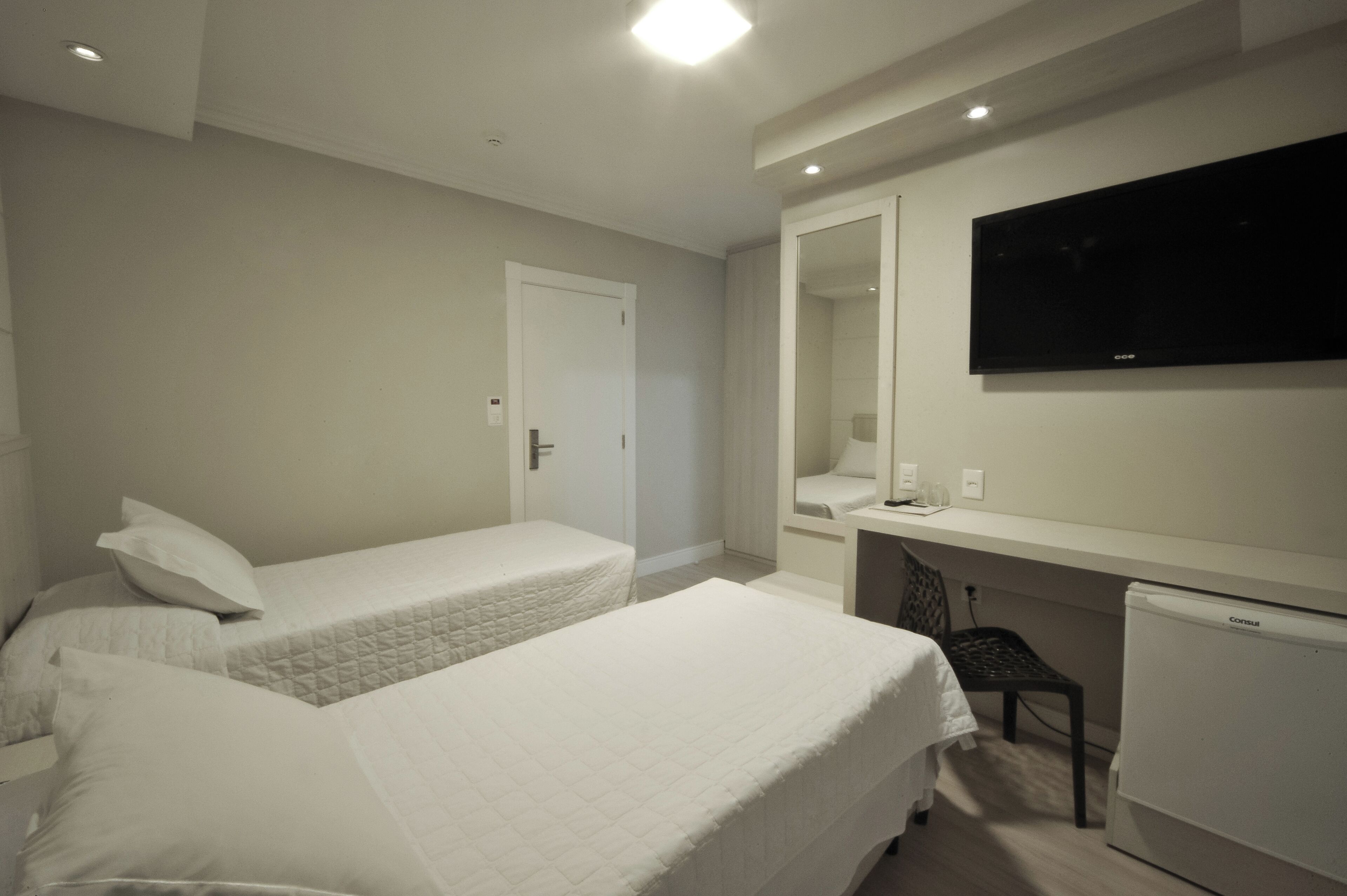 Habitación de hotel con dos camas individuales, un escritorio, una silla, un televisor y un refrigerador.