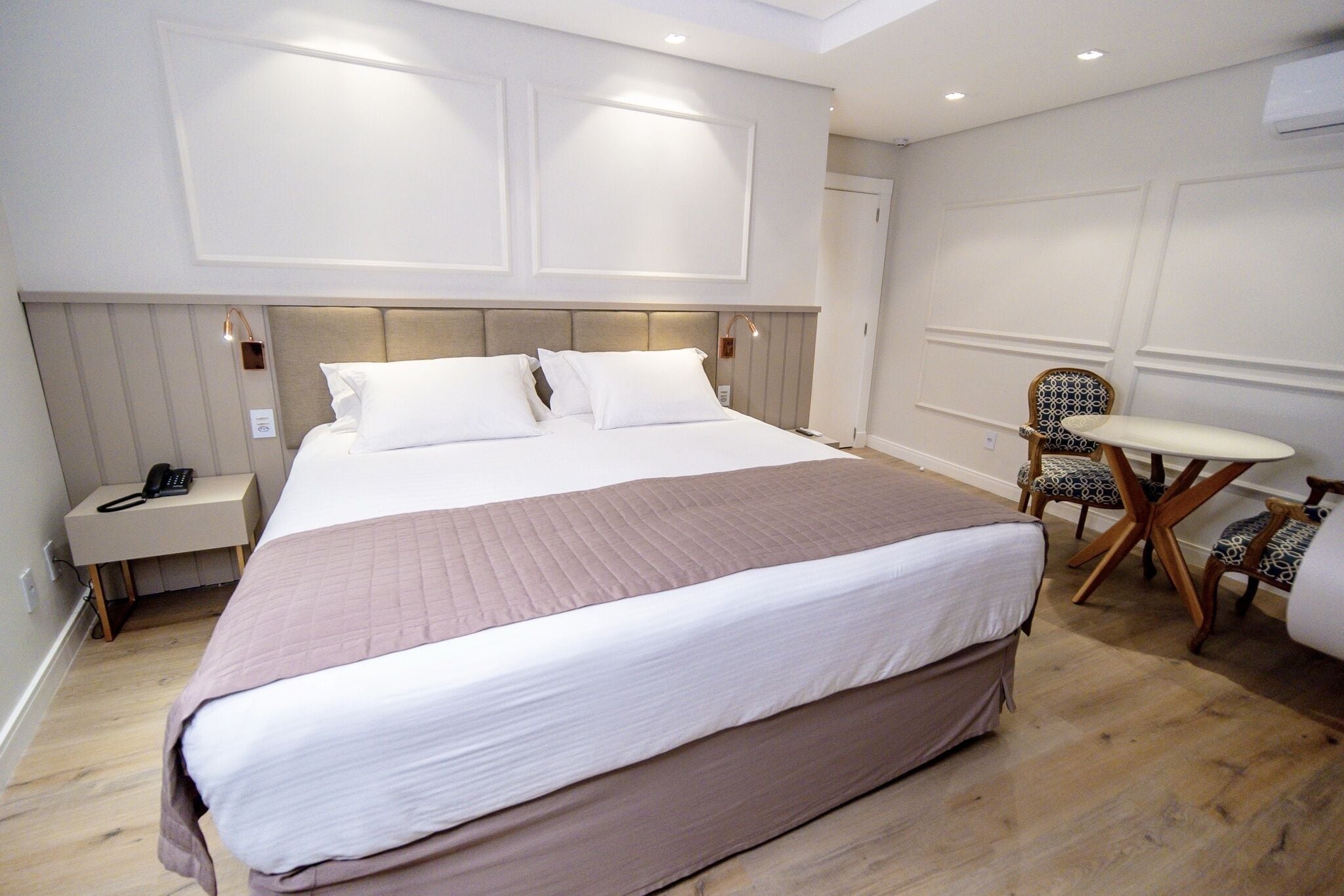 Quarto de hotel com cama grande, mesas de cabeceira, uma pequena mesa com cadeiras e telefone fixado na parede.