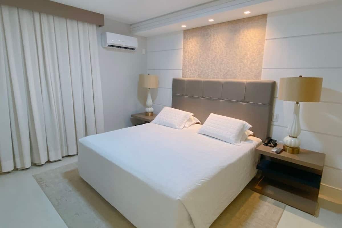 Una habitación de hotel moderna con una cama grande, dos mesitas de noche con lámparas, aire acondicionado de pared y un amplio ventanal con cortinas.