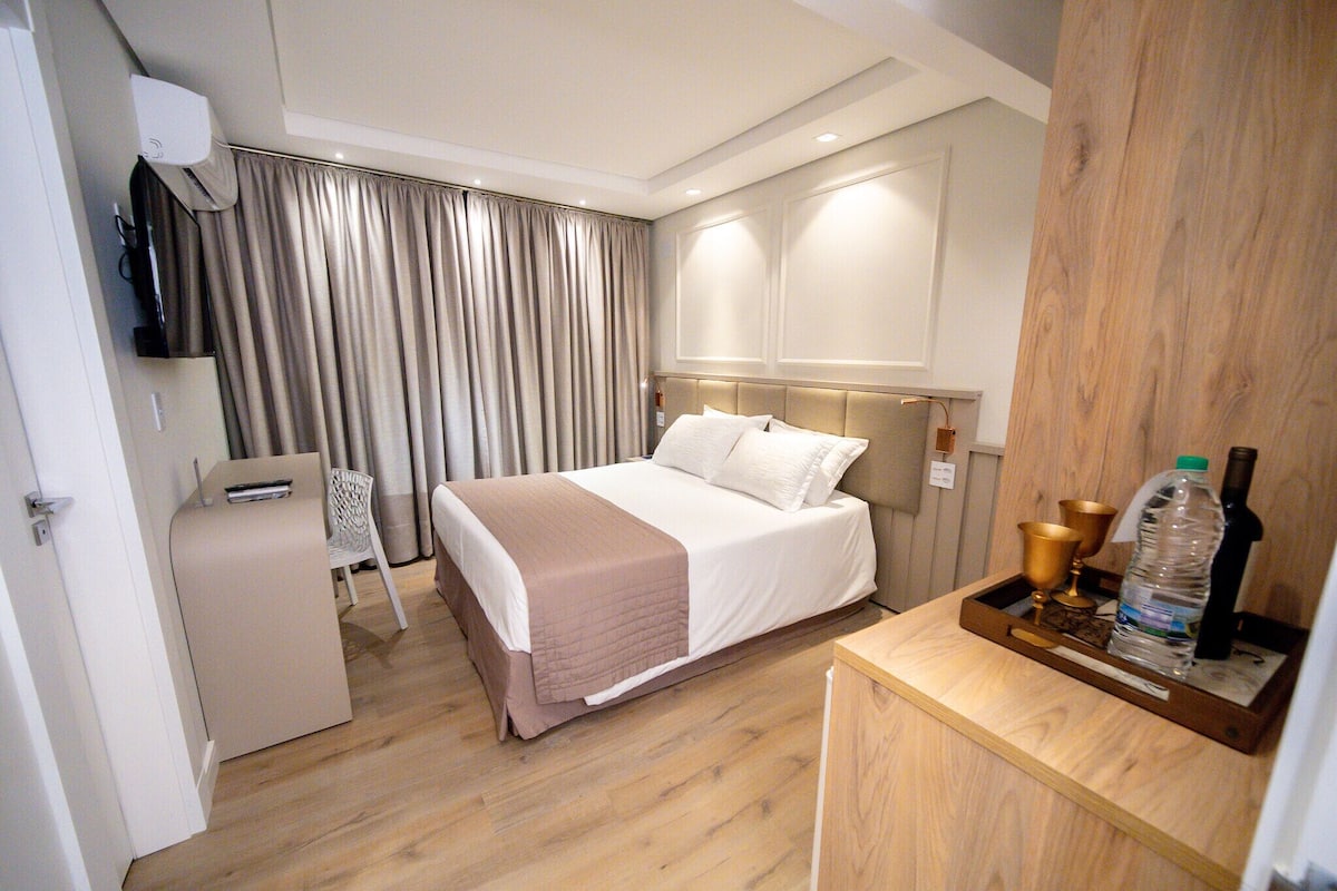 Una habitación de hotel con una cama, un escritorio con una botella y vasos, un televisor y una cortina.
