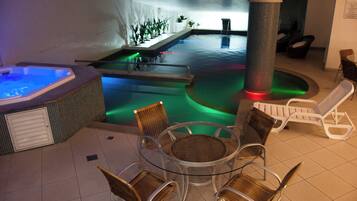 Una piscina interior con un jacuzzi, sillas de descanso y una mesa de vidrio rodeada de sillones de mimbre.