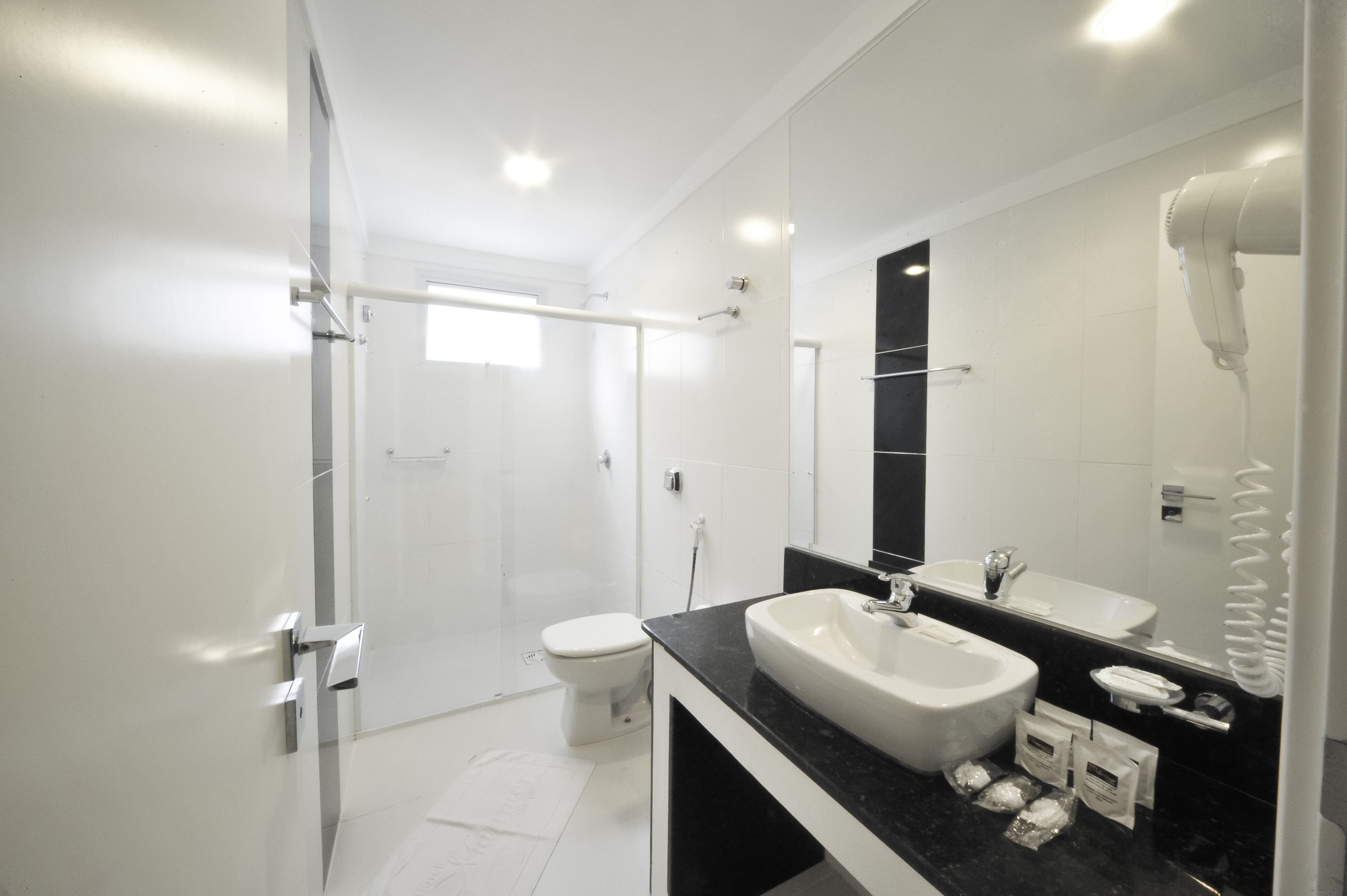 Ein modernes Badezimmer mit Duschkabine aus Glas, einer weißen Toilette, einer schwarzen Ablagefläche mit Waschbecken und einem an der Wand befestigten Föhn.