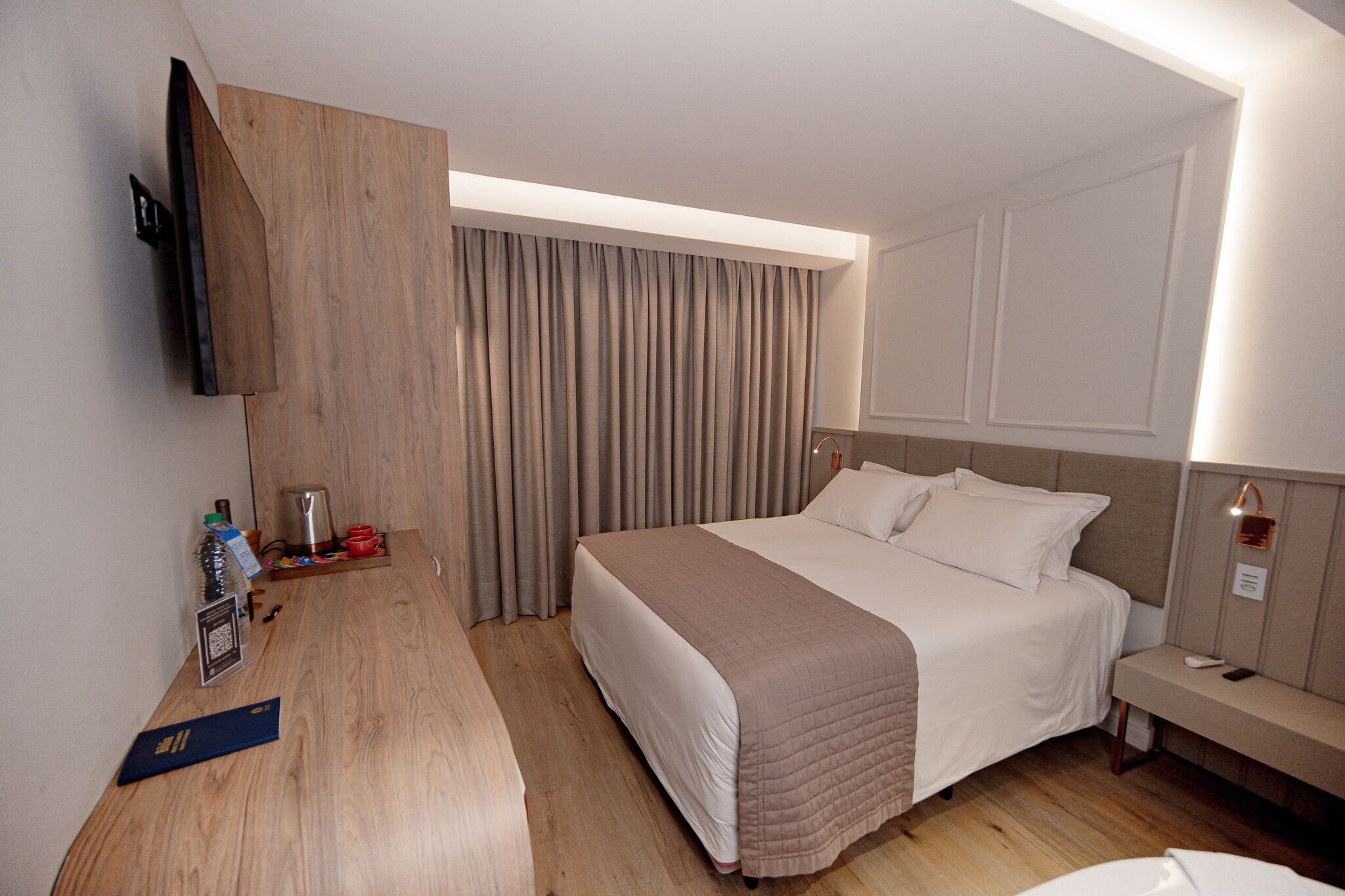 A hotel room with a large bed, a bedside table, a TV mounted on the wall, and a wooden headboard.