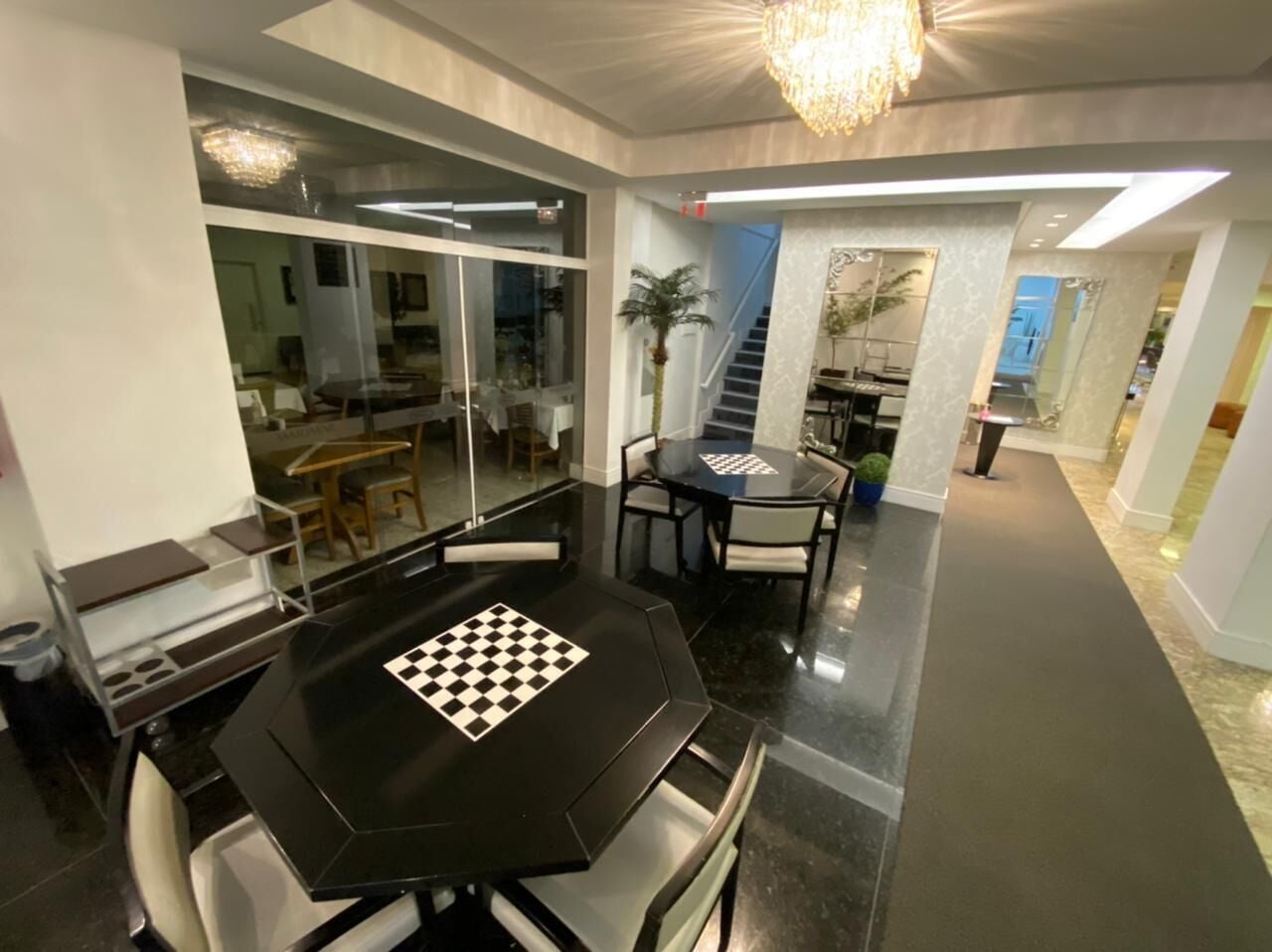 A modern dining area with a black table featuring a chessboard, glass walls, and a chandelier.