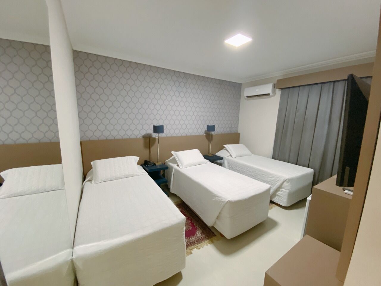 Quarto de hotel com três camas de solteiro, uma pequena escrivaninha, um criado-mudo e uma televisão.