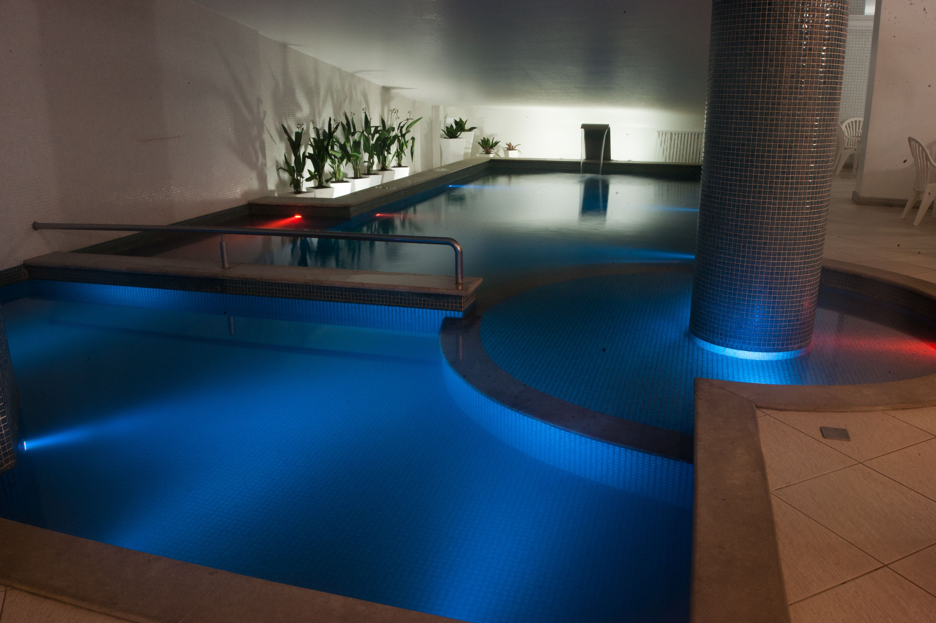 A swimming pool with a blue-lit edge, a tiled floor, and potted plants along the wall.