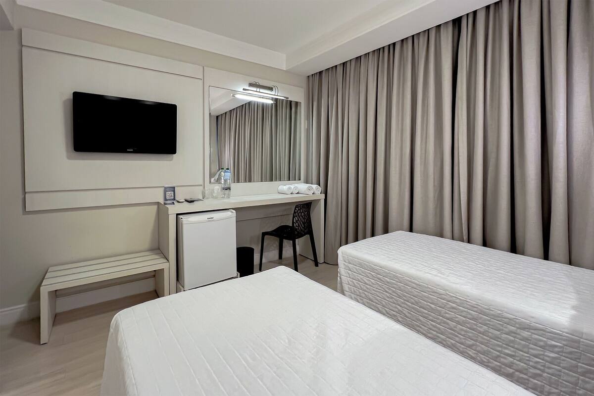 A hotel room with a bed, a desk with a computer, a television, and a large window with curtains.