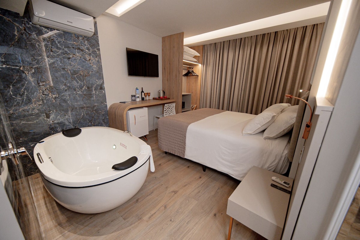 Una habitación de hotel moderna con una bañera grande, una cama con ropa blanca, un escritorio con computadora y un televisor montado en la pared.