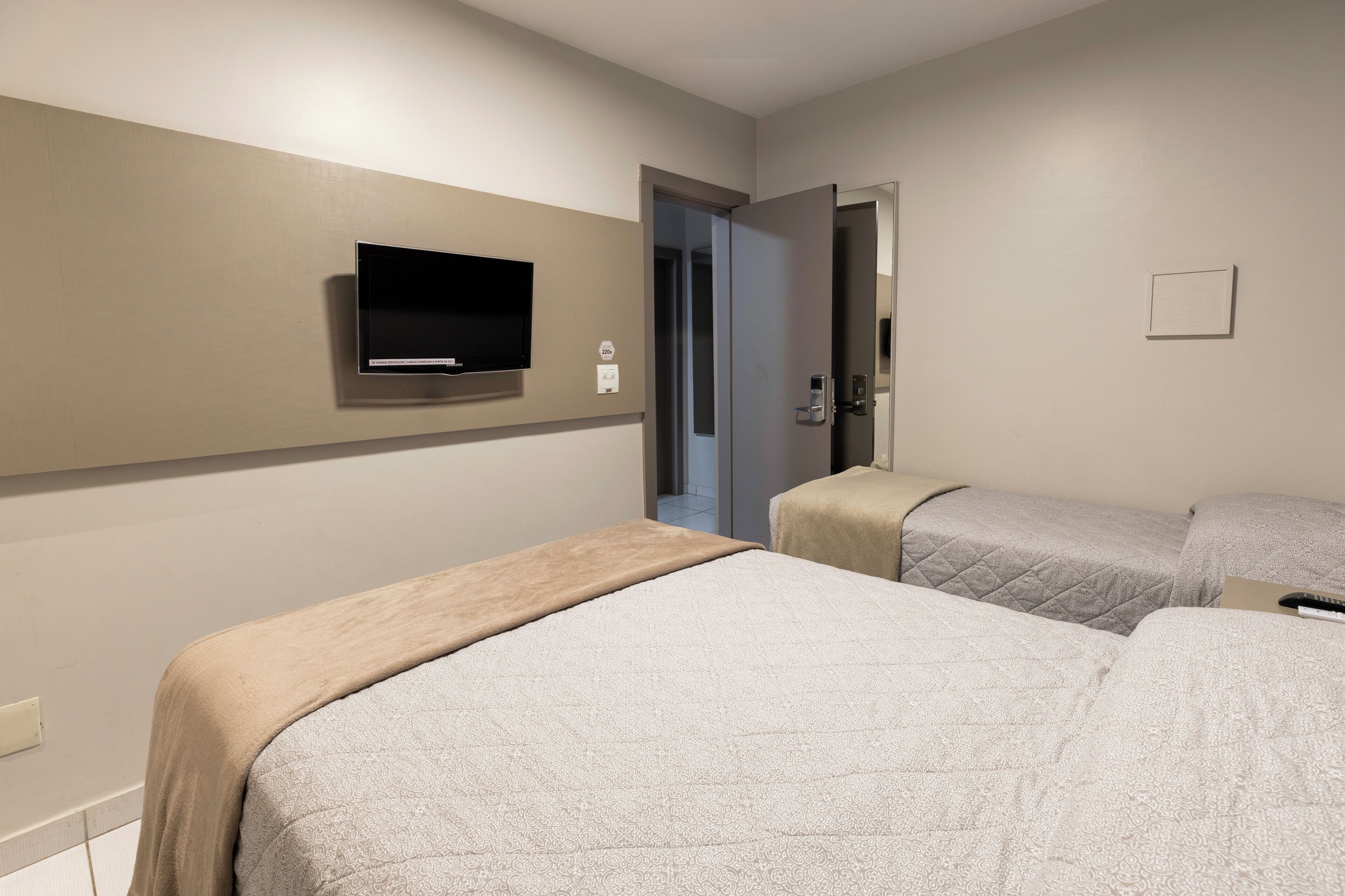Habitación de hotel con dos camas, un televisor montado en la pared y una puerta que da a otra habitación.