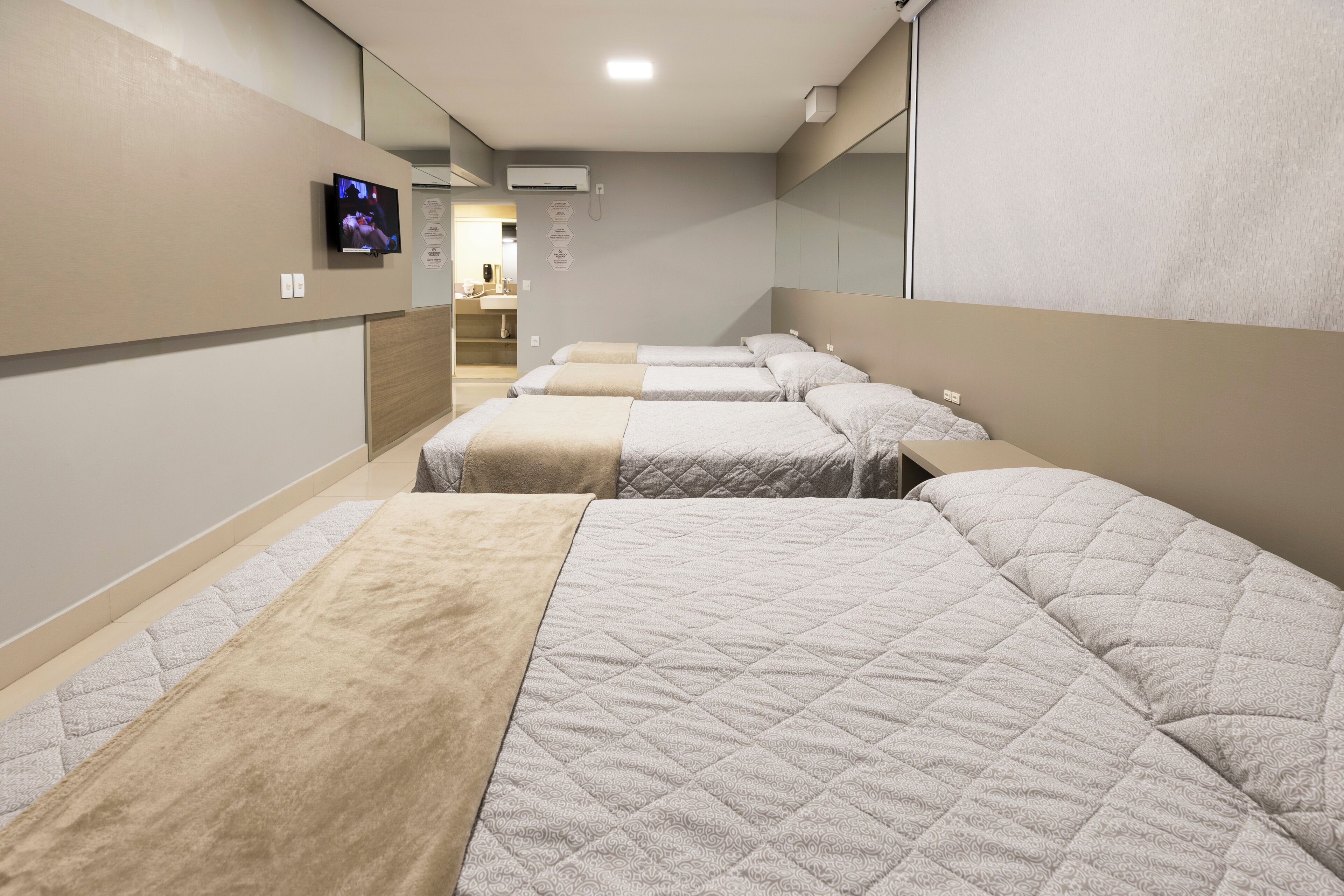 A hotel room with four beds arranged in a row, a television mounted on the wall, and a bathroom visible through an open door.