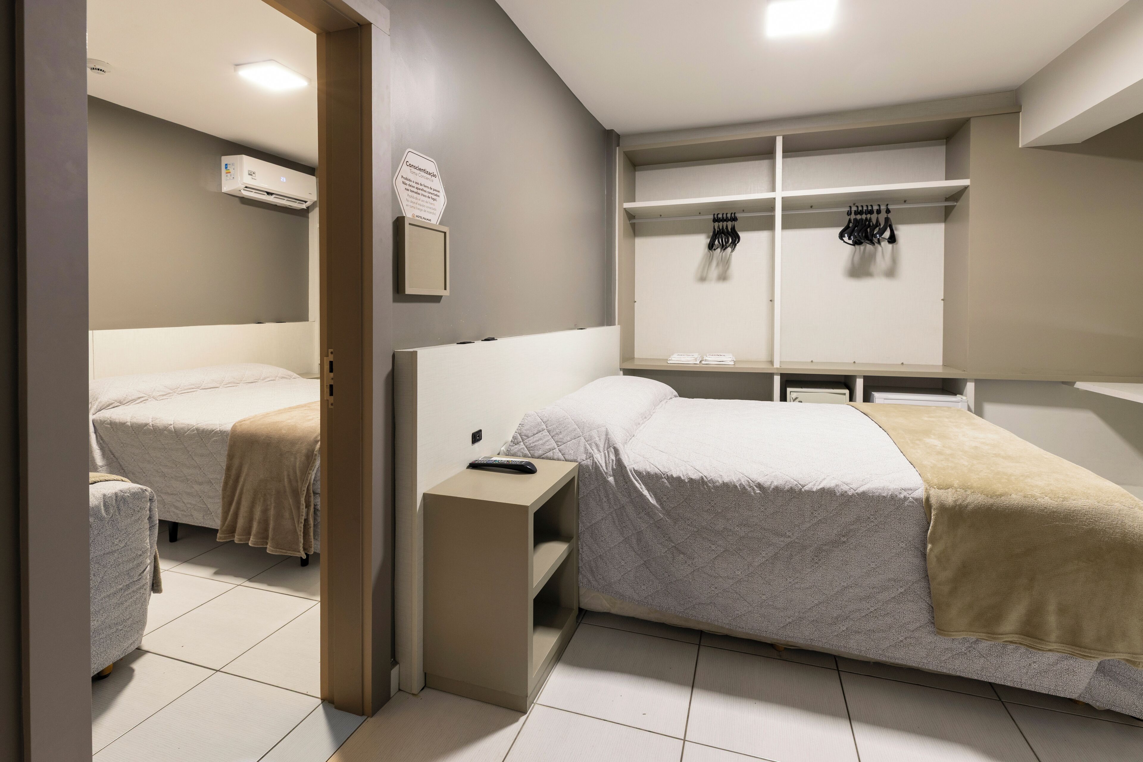 Habitación de hotel con dos camas, una mesita de noche pequeña, un armario con ropa colgada y un aire acondicionado de pared.