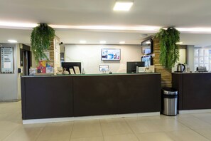 A reception desk with a digital screen, a trash bin, and a wall-mounted TV. - Hotel Palmas Executivo (Balneário Camboriú)