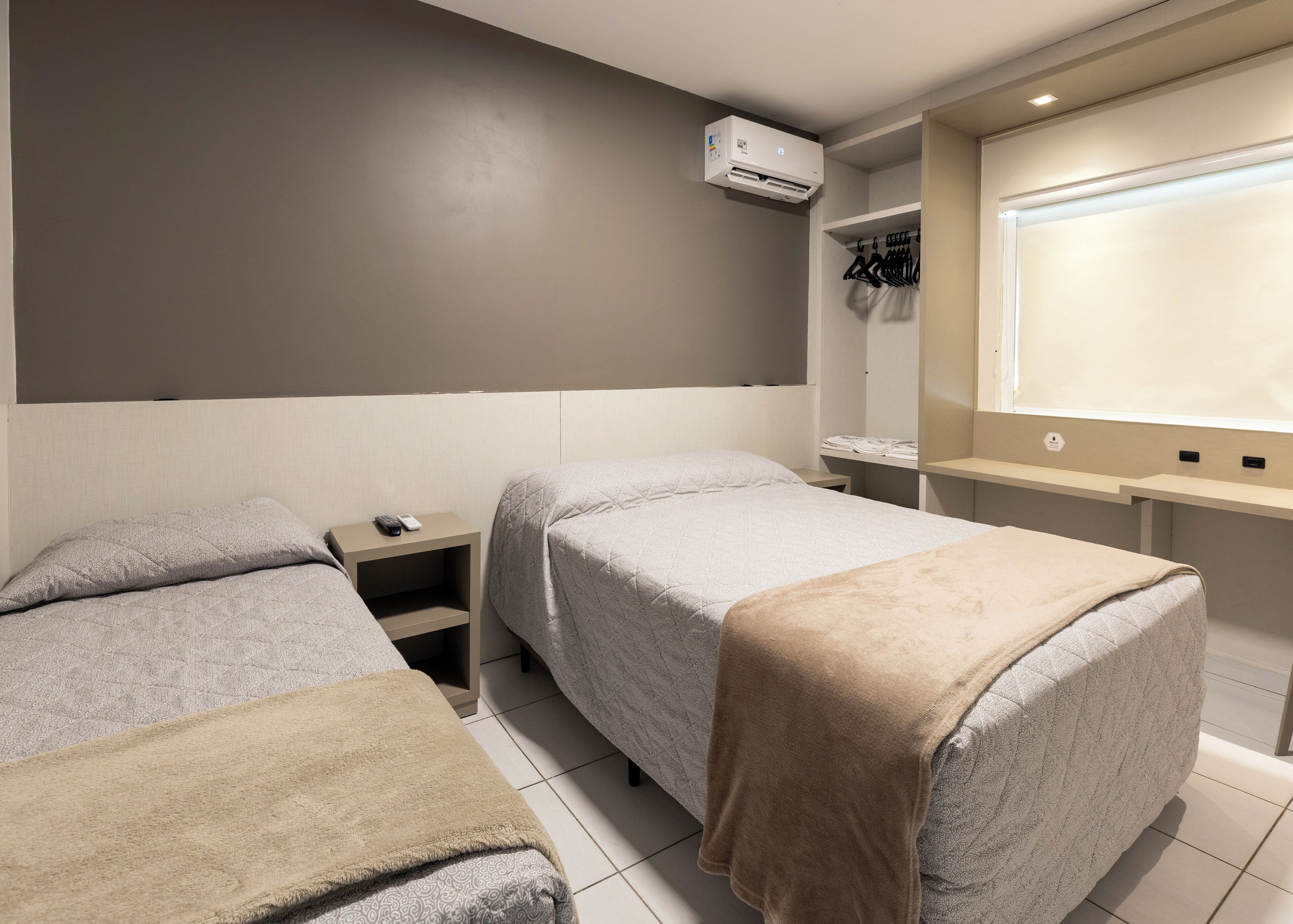 A hotel room with two beds, a bedside table with a remote control, a desk with a computer, and a wardrobe with hanging clothes.