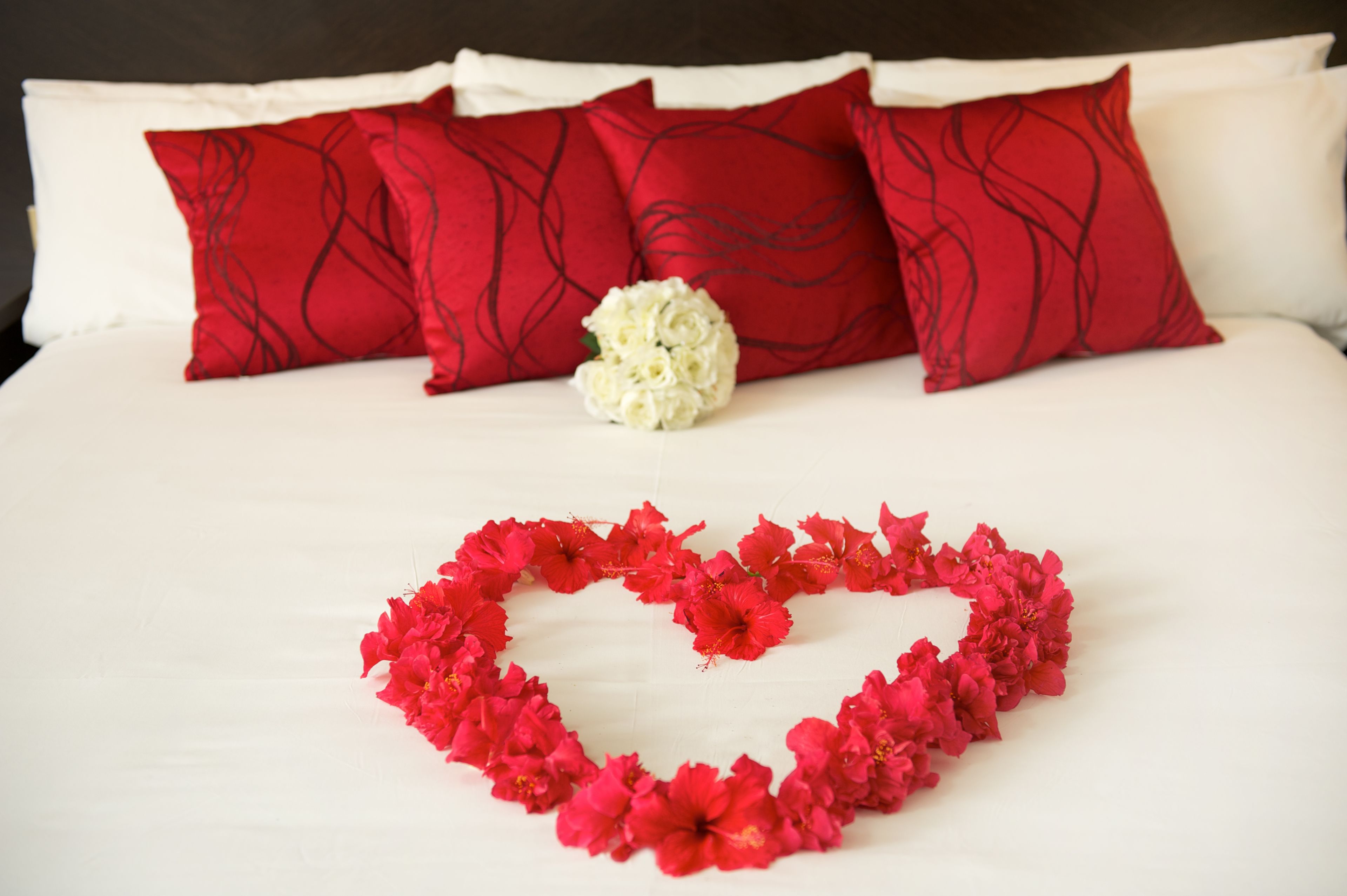 A bed with red and white pillows, a white flower, and a red flower heart shape on the bed.