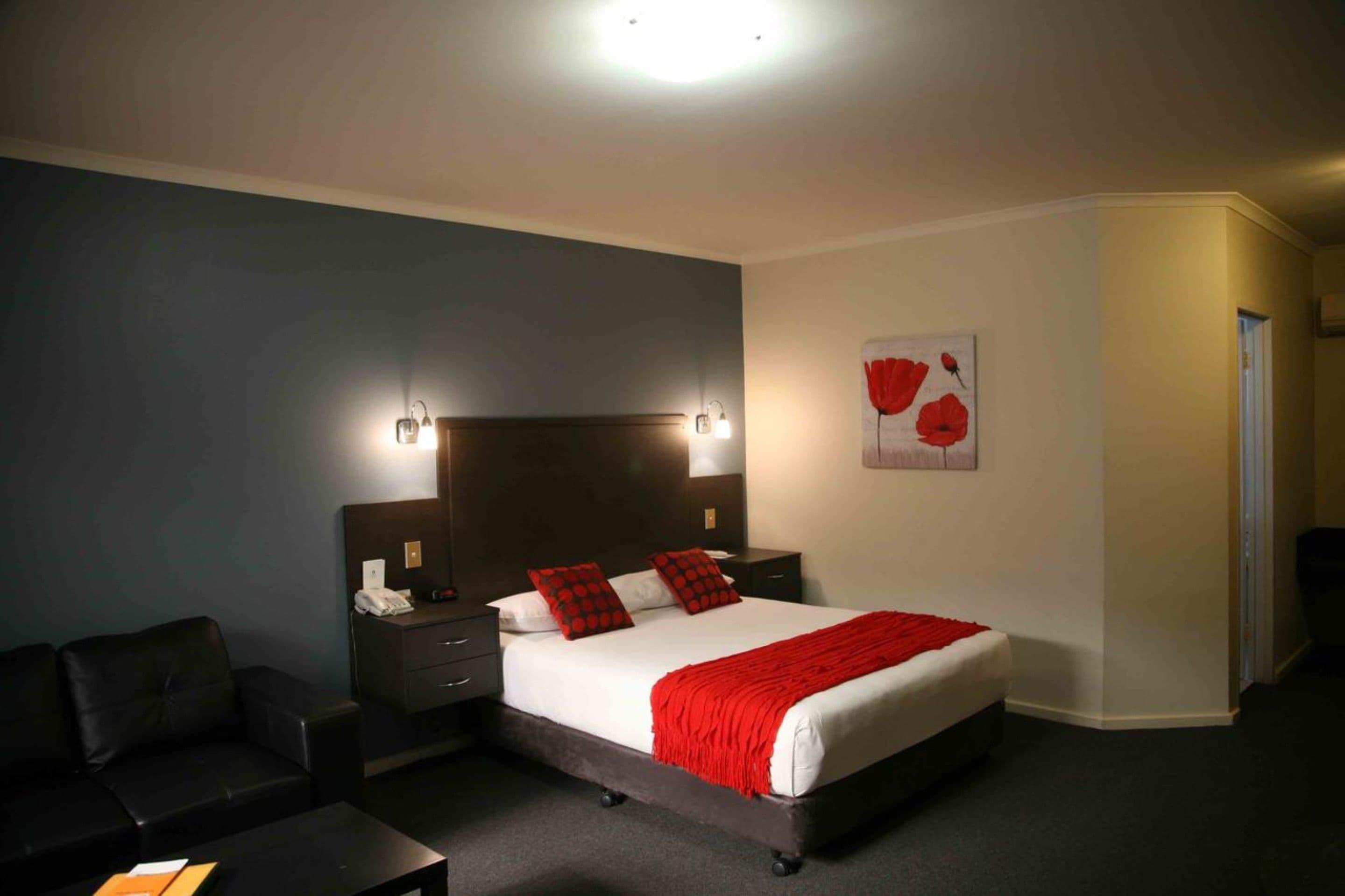 A hotel room with a large bed, two bedside tables, a nightstand, a black leather sofa, and a red flower painting on the wall.