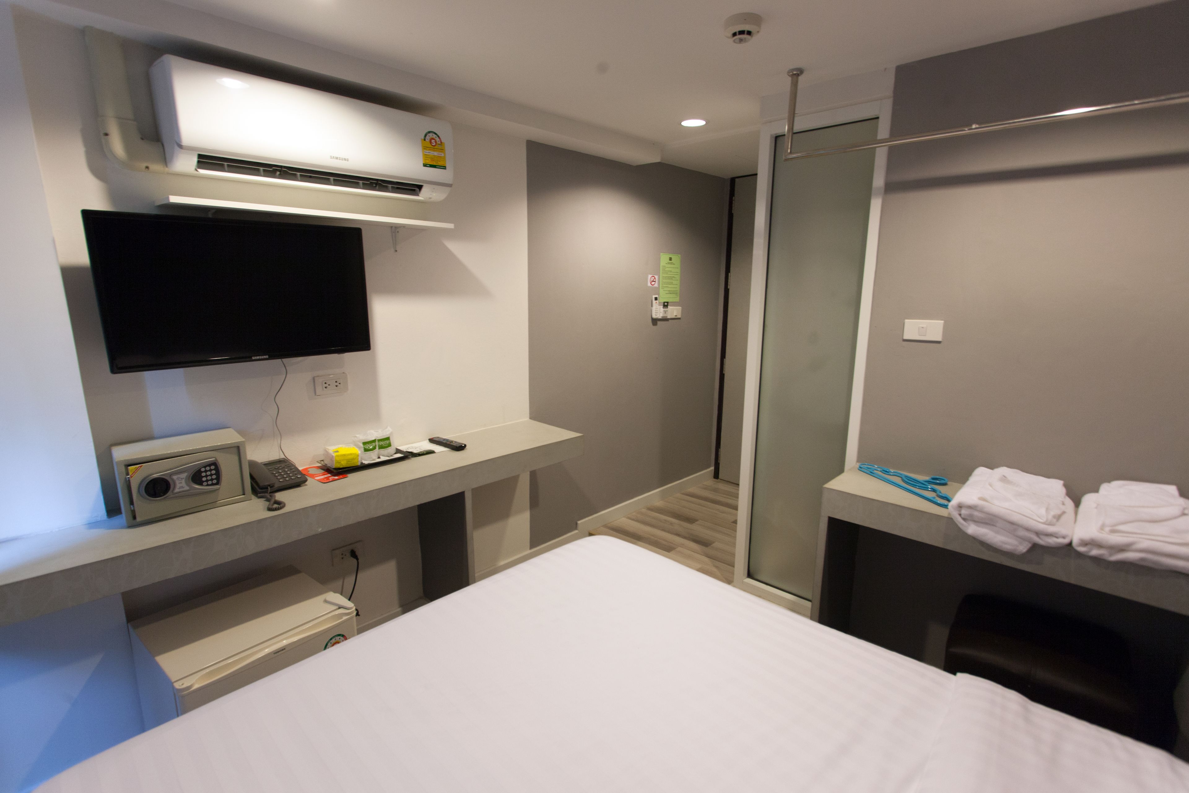 A hotel room with a bed, a flat-screen TV, a desk with a radio and remote controls, a shower area, and a towel rack.