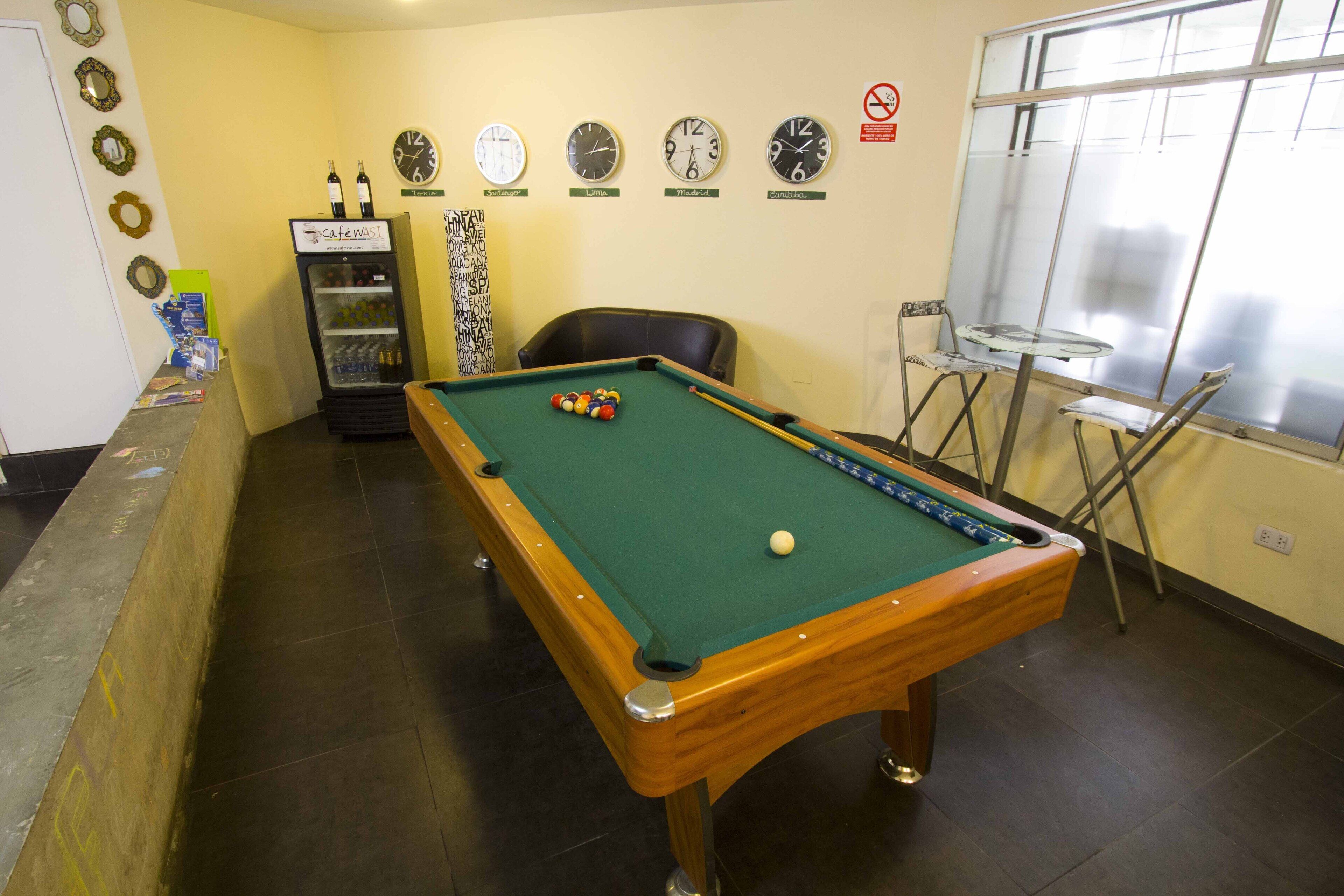 Una mesa de billar con superficie de fieltro verde, una cola de billar y bolas de billar. Hay relojes en la pared y un refrigerador con botellas.