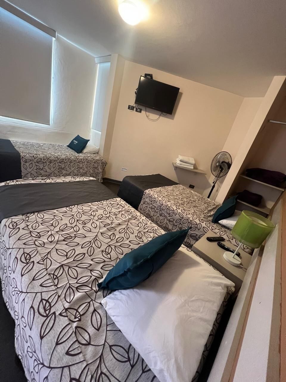 A hotel room with two beds, a TV, a fan, and a window with blinds.