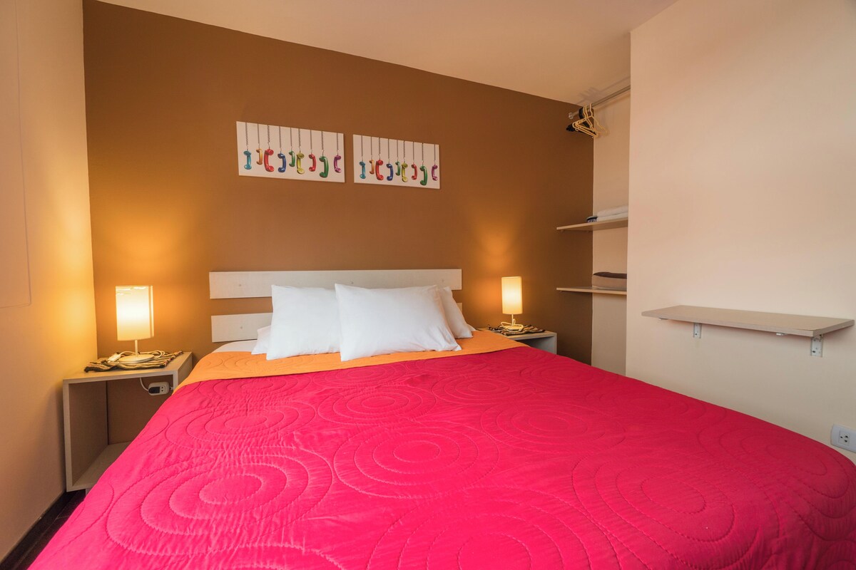 A hotel room with a large bed, two bedside lamps, a nightstand, a wall-mounted shelf, and a wall art piece.
