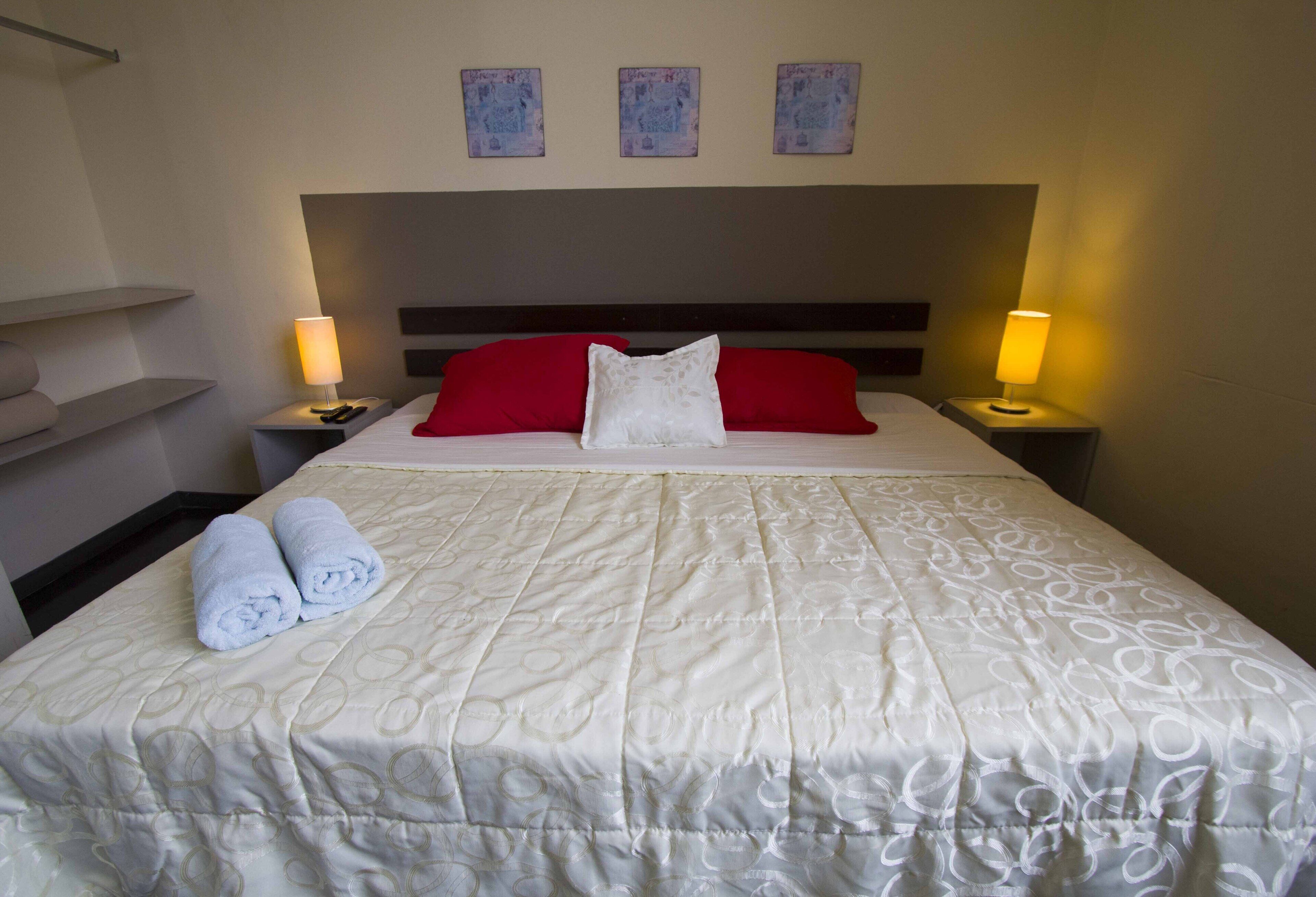 A neatly made bed with a white bedspread, red and white pillows, and two rolled towels on the side. There are two bedside lamps, a headboard, and three framed pictures on the wall.
