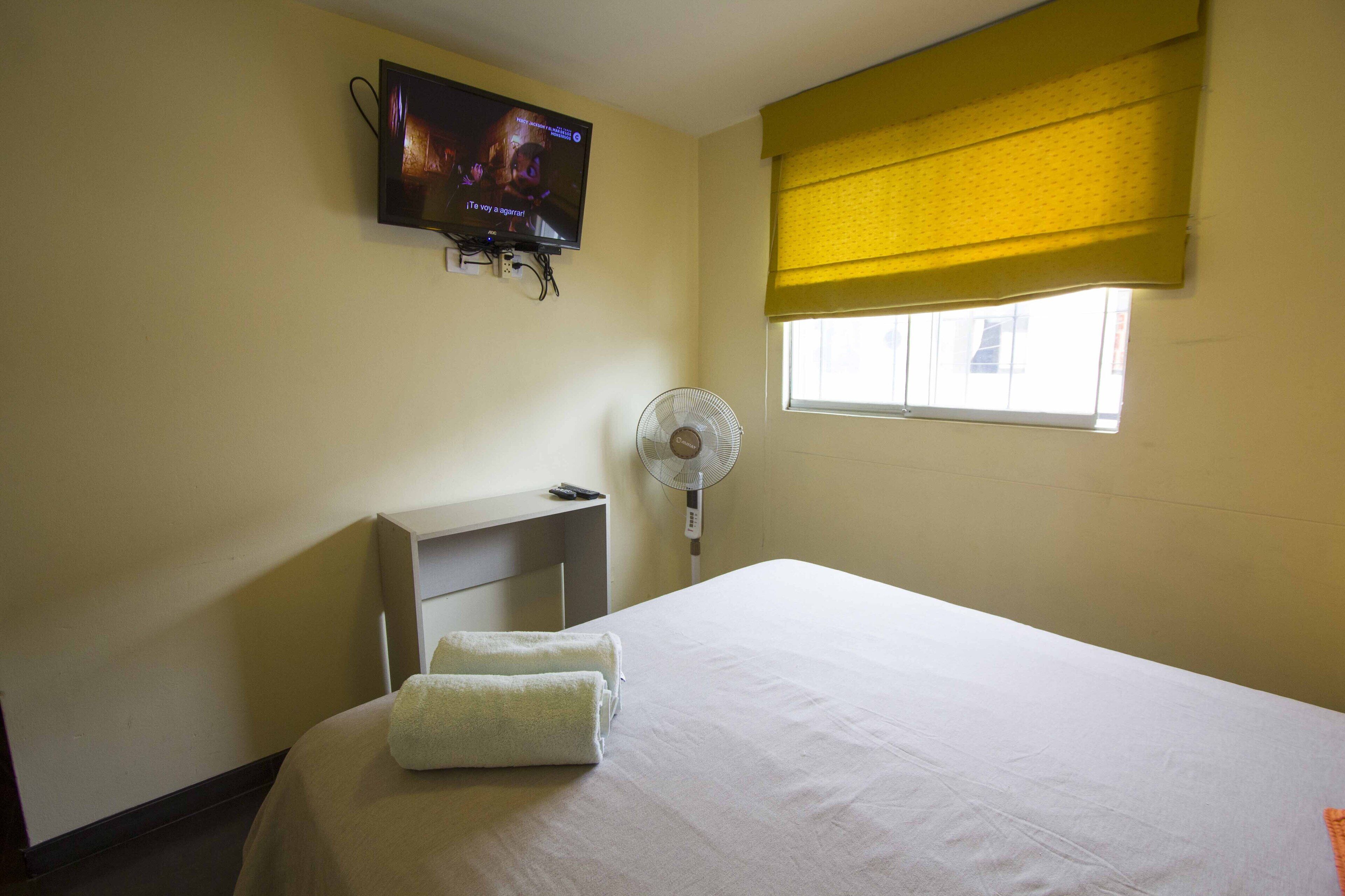 Hotelkamer met een bed, een aan de muur bevestigde televisie, een nachtkastje, een ventilator en een raam met gele zonwering.