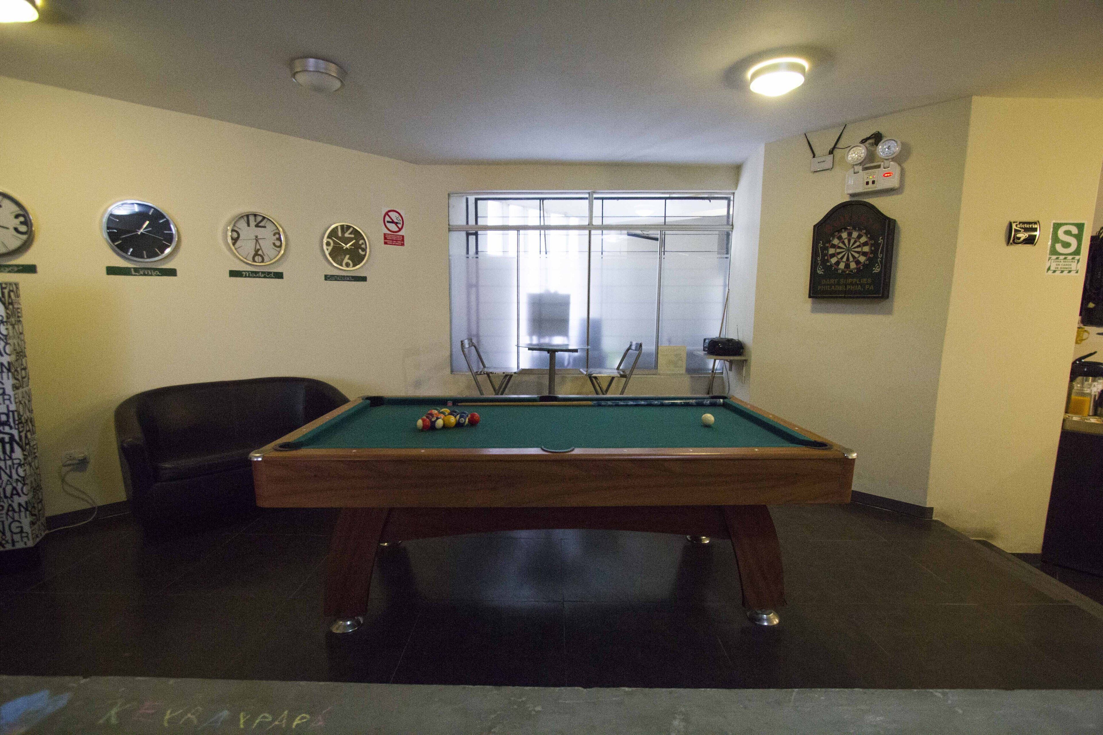 A pool table with balls, a clock, and a dartboard in a room with a window and a chair.