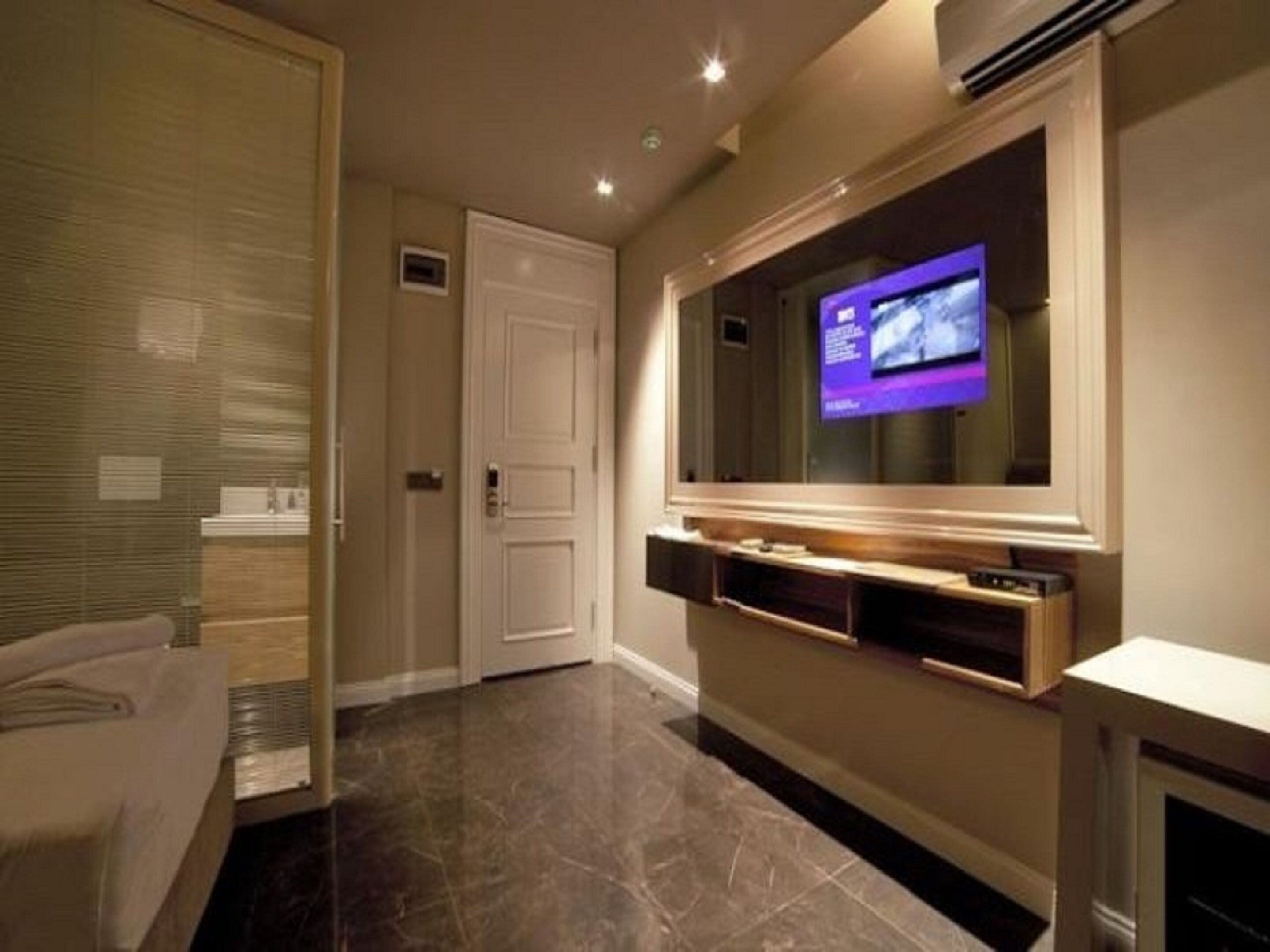 A modern hotel room with a large flat-screen TV mounted on the wall, a marble-tiled floor, and a white door.