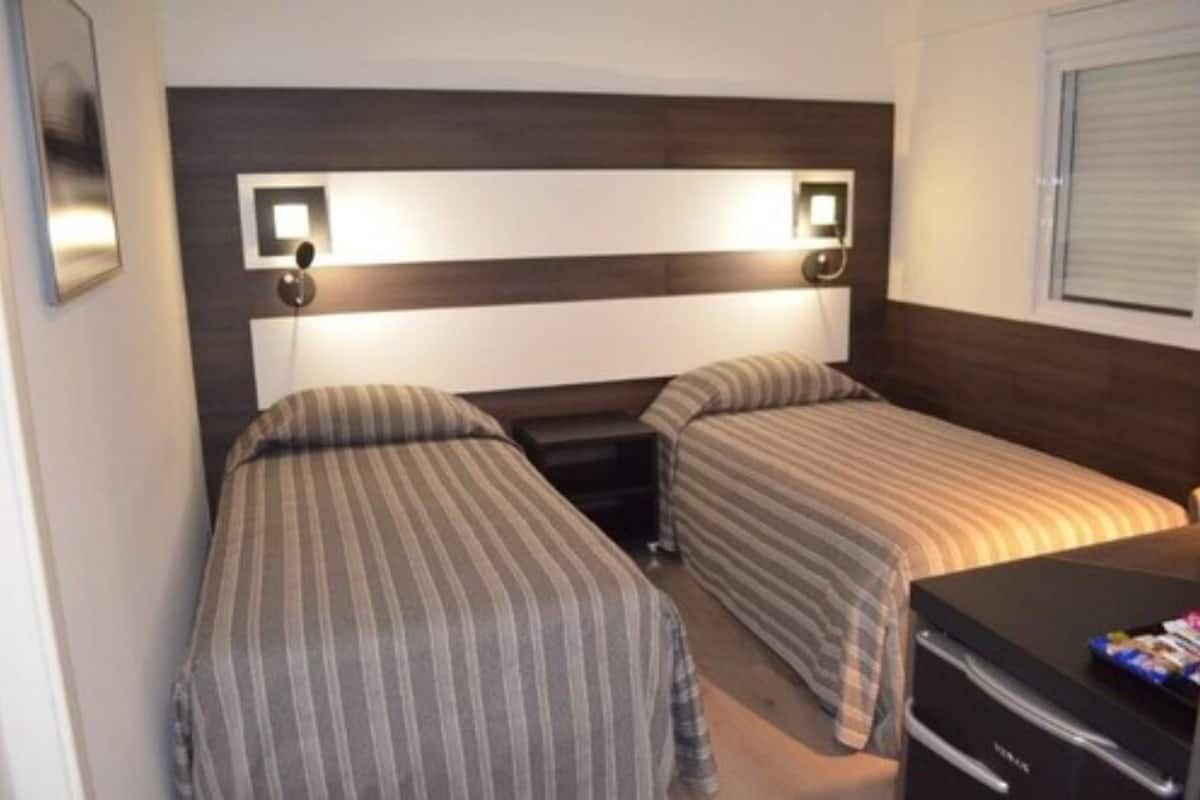 A room with two single beds, a desk with a computer, and a window with blinds.