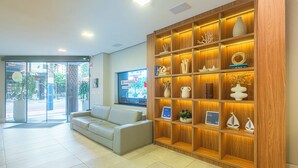 A reception area with a light-colored sofa, a wooden display shelf with various decorative items, and a large window with a view of the street. - Hotel Melo (Balneário Camboriú)