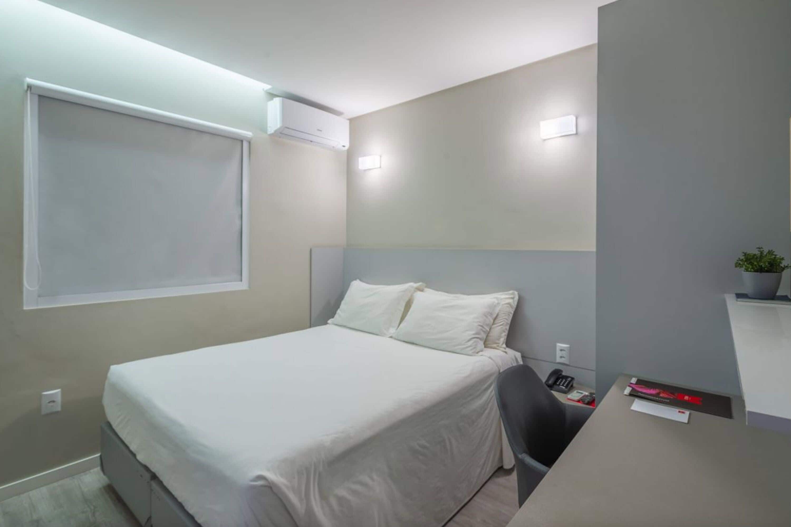Une chambre à coucher avec un lit, un bureau et une chaise. Il y a une fenêtre avec un store, un appareil de climatisation et une petite plante sur le bureau.