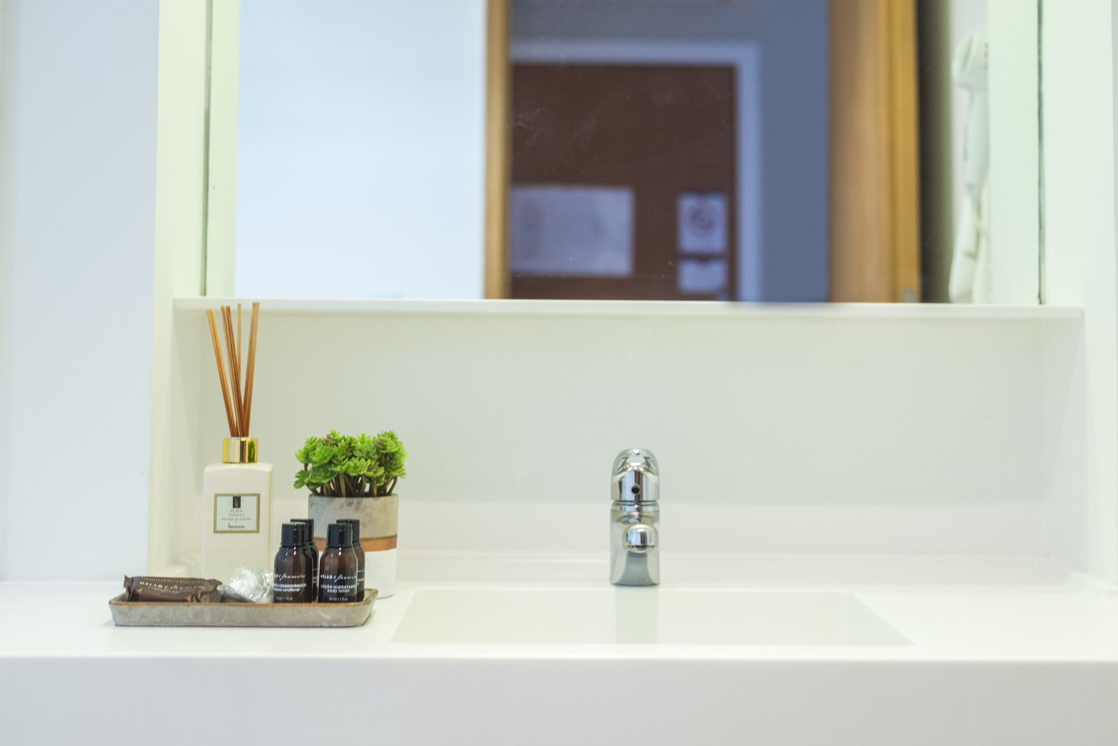 Ein Badezimmer mit einem weißen Waschbecken, einem Spiegel und einer kleinen Pflanze. Auf dem Waschbecken befinden sich außerdem zwei Flaschen und eine Schere.