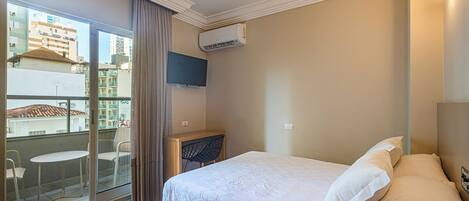 A hotel room with a bed, a TV, a balcony with chairs, and an air conditioning unit.