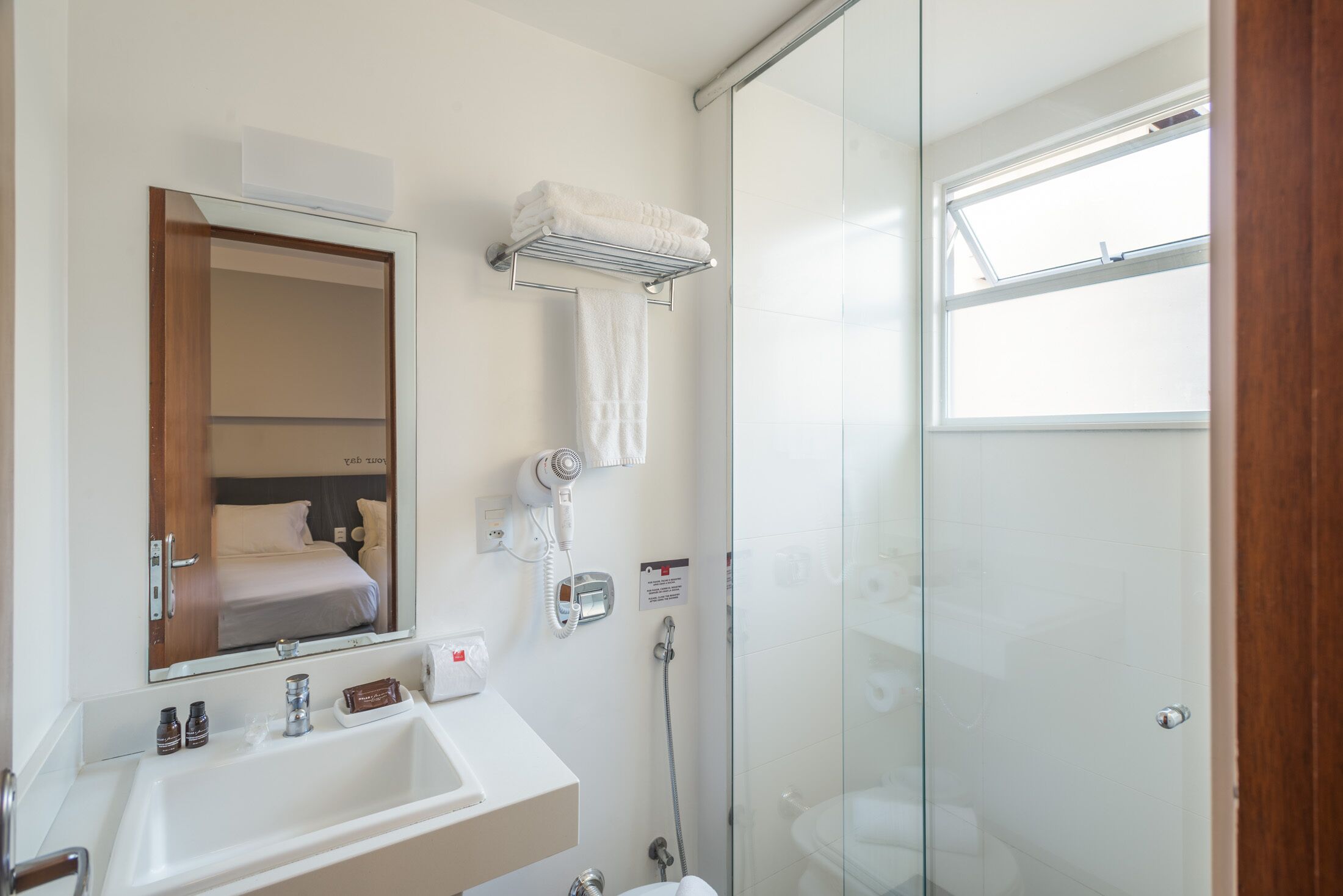 Une salle de bain moderne avec une douche à parois de verre, un lavabo avec des articles de toilette et un miroir qui reflète un lit.