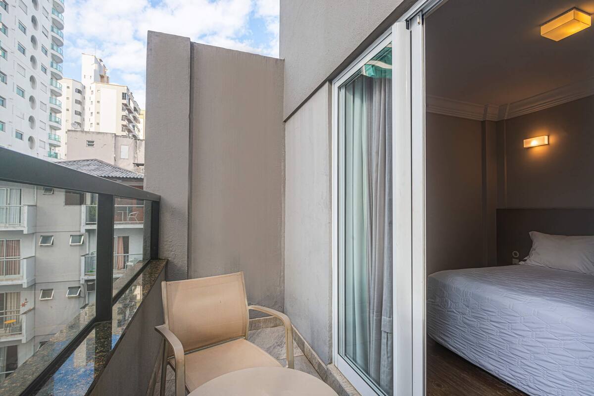 A hotel room with a bed, a chair, a balcony with a view of other buildings, and a window with curtains.
