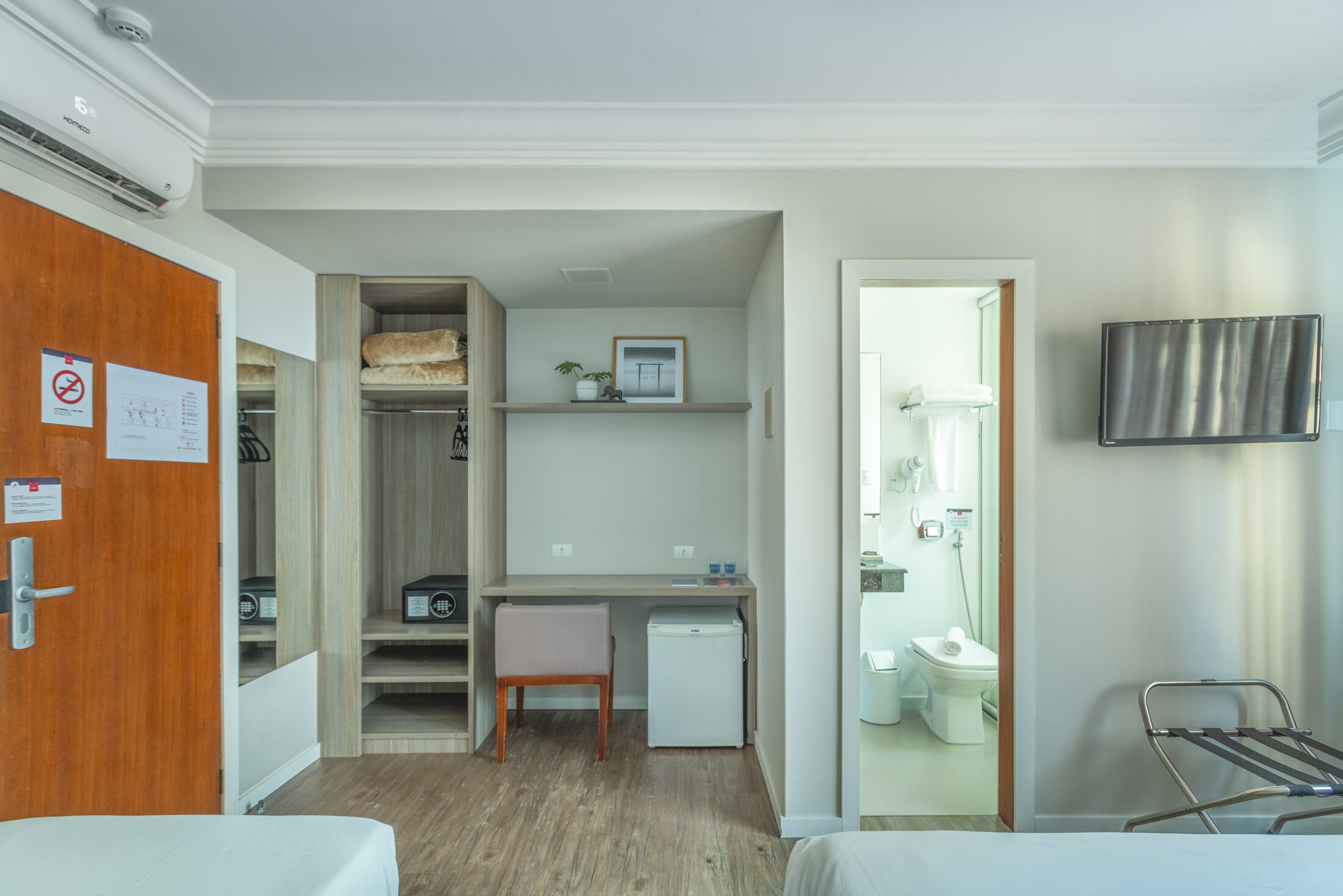 Une chambre d’hôtel de taille réduite, comprenant un lit, un bureau, une chaise, un petit réfrigérateur, une télévision et une salle de bain avec des toilettes et une douche.