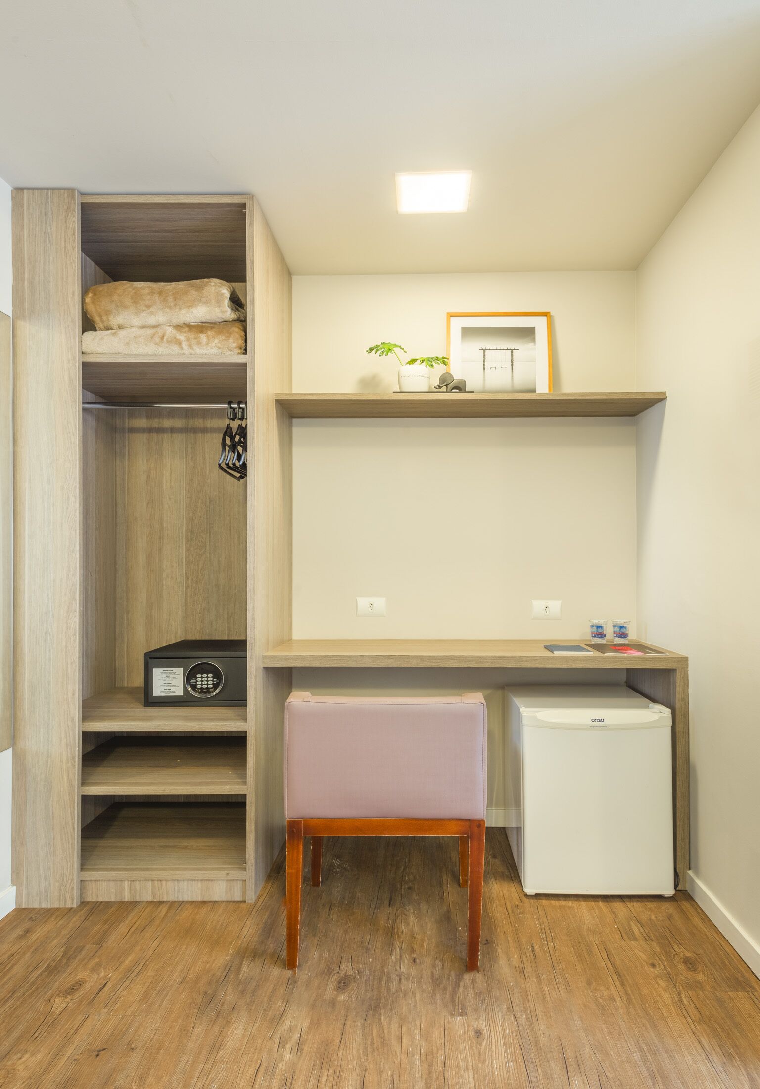 A compact room with a wooden desk, a small refrigerator, a chair, and a shelving unit with a radio and folded blankets.