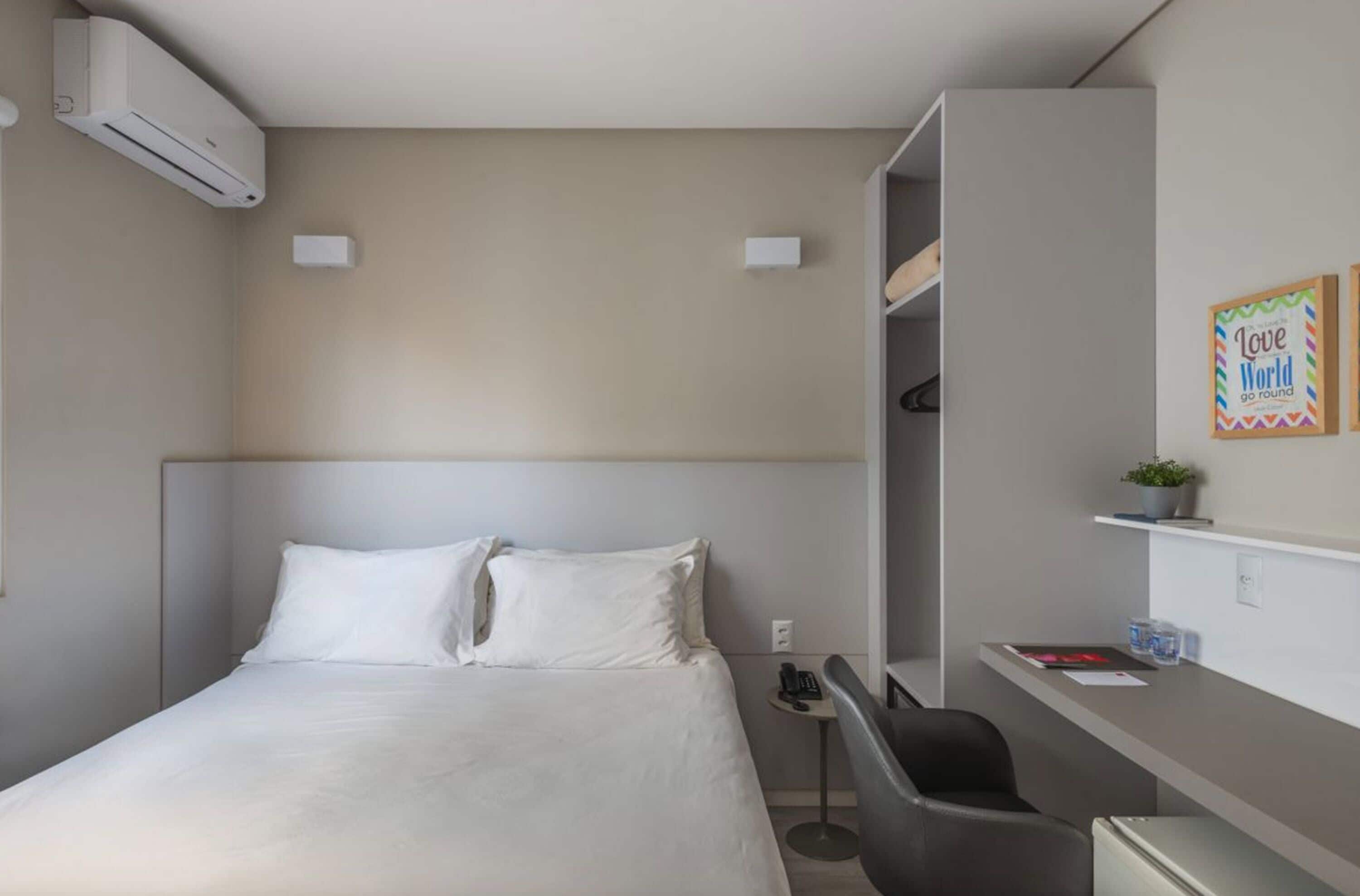 A hotel room with a bed, a desk, a chair, an air conditioner, and a shelf with a plant and a sign.