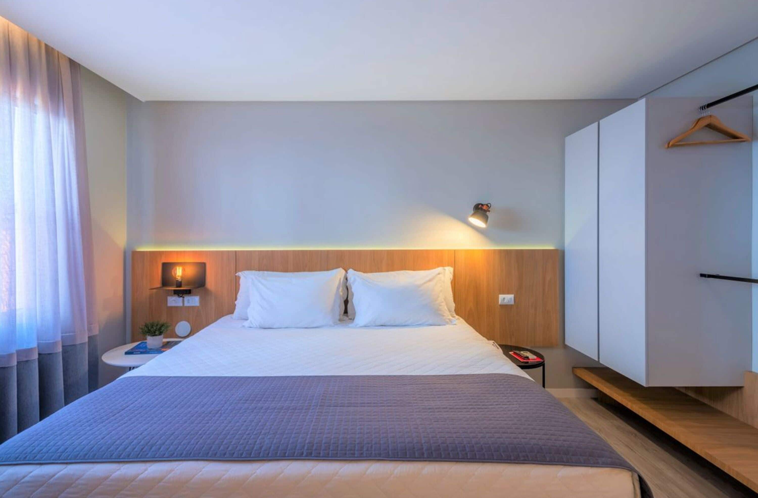 A modern hotel room with a large bed, a bedside table with a lamp, a wardrobe, and a hanging coat rack.