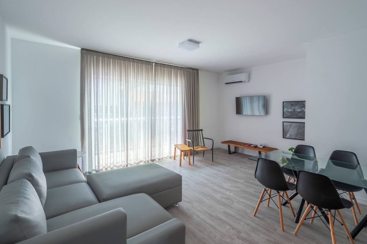 A modern living room with a grey sectional sofa, a glass dining table with black chairs, a wooden bench, and a TV mounted on the wall.