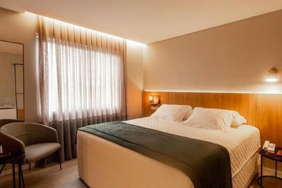 Une chambre d’hôtel avec un lit, une chaise, un bureau et une fenêtre avec des rideaux.