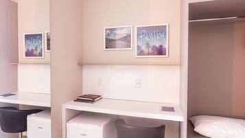 Habitación de hotel con una cama litera, un escritorio con computadora, una silla y un refrigerador pequeño. Hay cuadros en la pared.
