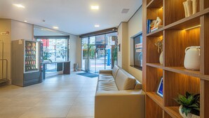 A hotel lobby with a reception desk, a vending machine, a seating area, and a wooden shelving unit with decorative items. - Hotel Melo (Balneário Camboriú)
