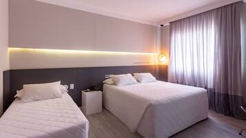A hotel room with two beds, a bedside table with a lamp, and a large window with curtains.