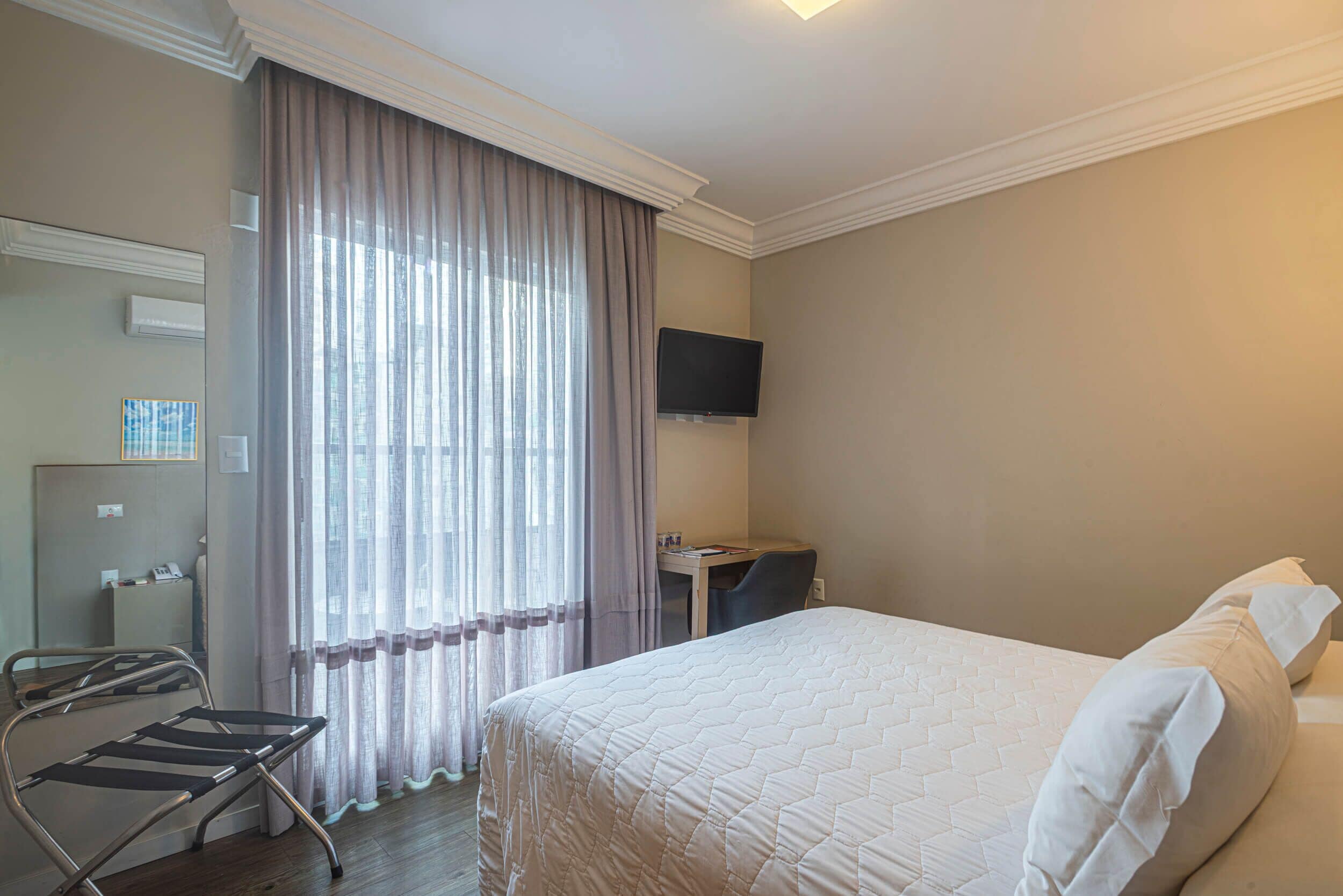 Habitación de hotel con cama, escritorio, silla, televisor y ventana con cortinas.