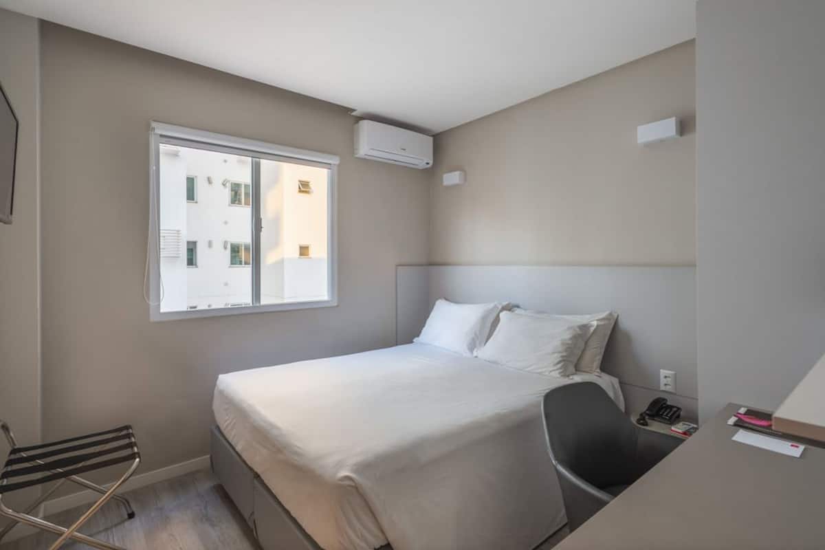A hotel room with a bed, a chair, a desk, a window, and an air conditioning unit.