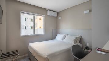 Quarto de hotel com cama, cadeira, escrivaninha, janela e ar condicionado.