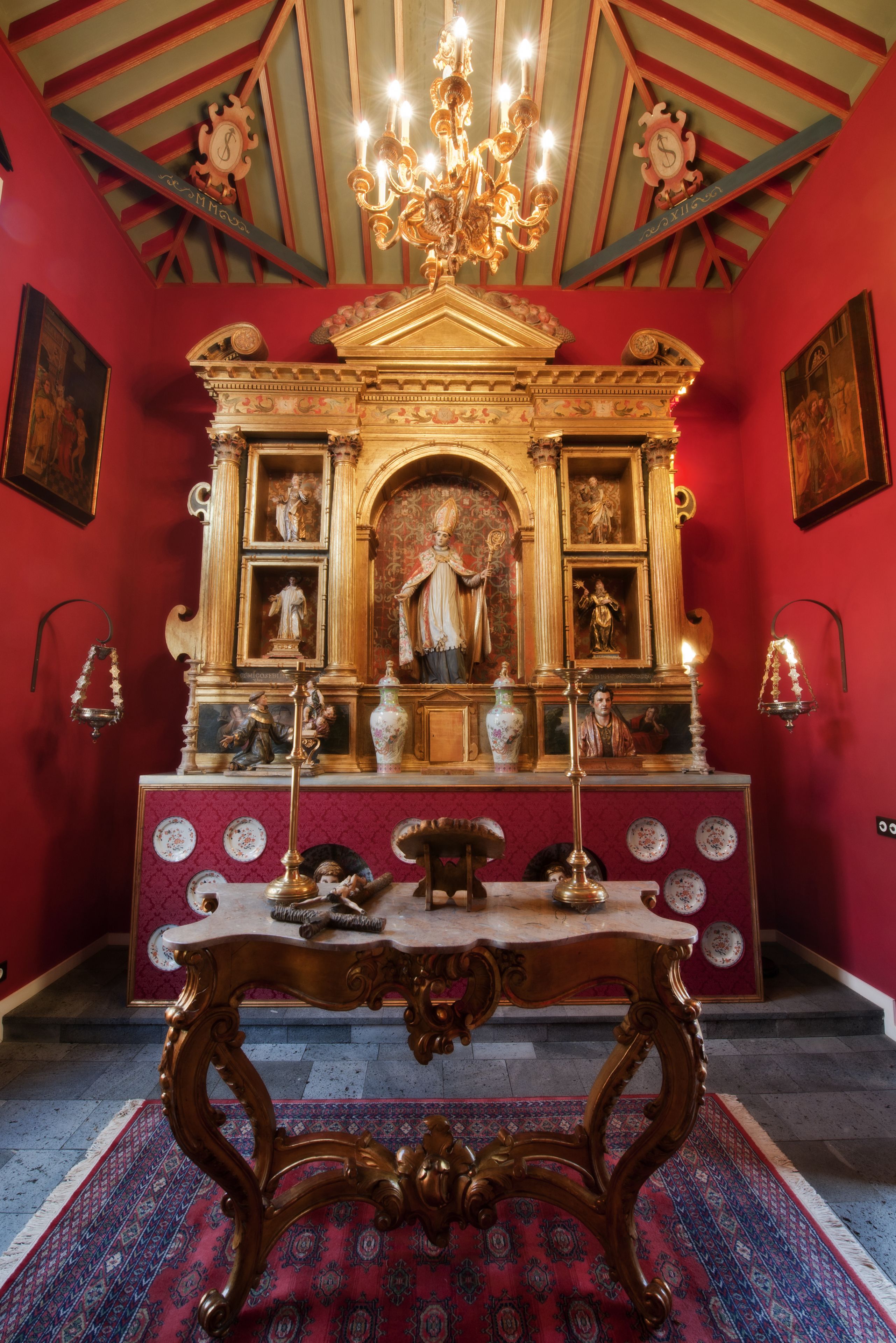 Une pièce avec un mur rouge, un lustre, une table aux pieds décorés et une statue religieuse.