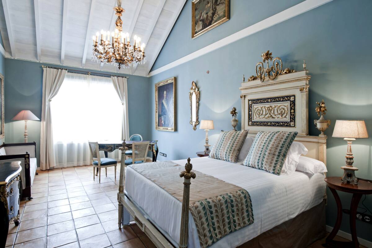 A bedroom with a bed, a chair, a desk, a chandelier, and a window with curtains.