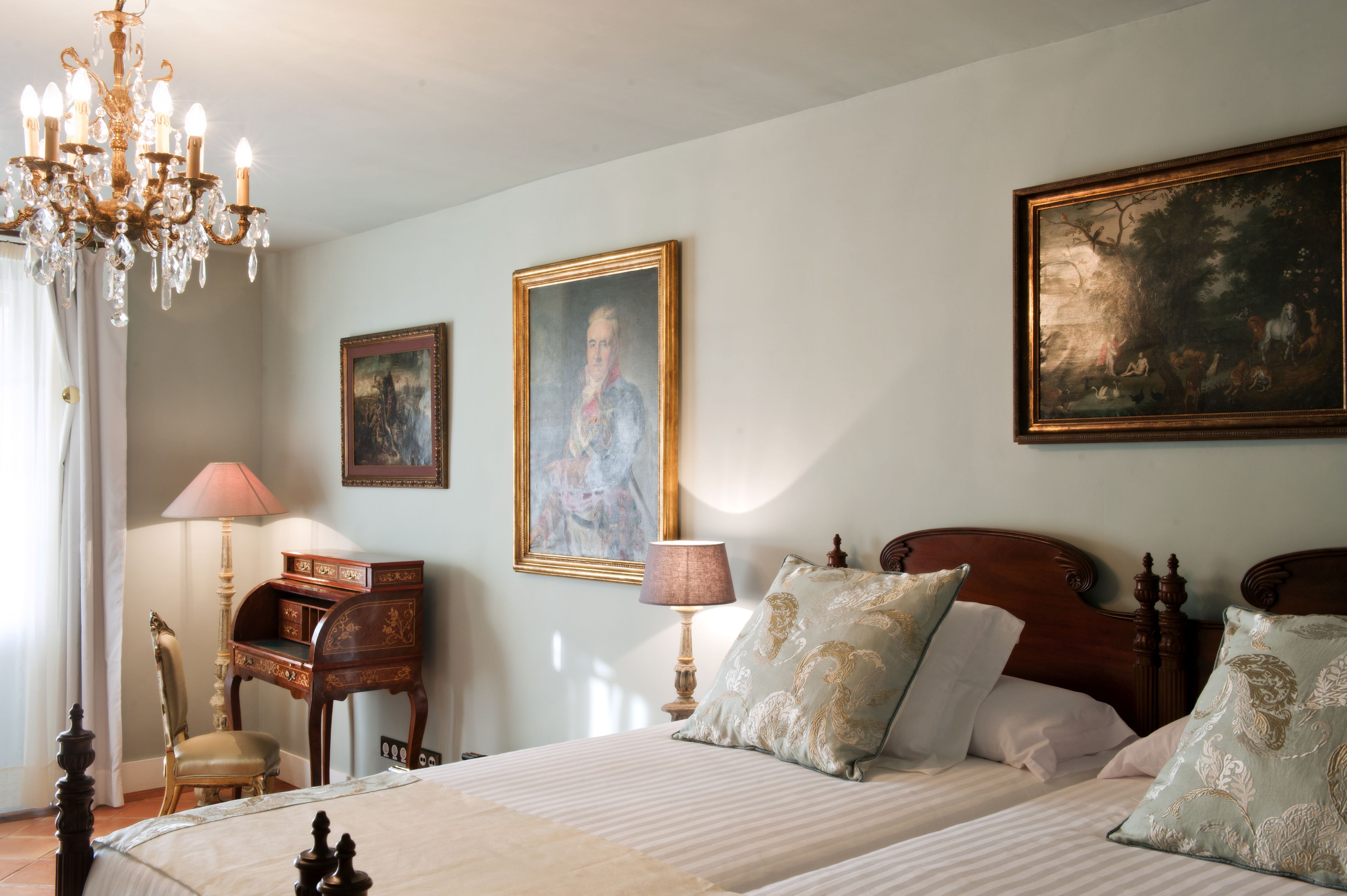 Quarto com uma cama grande, mesa de cabeceira de madeira, escrivaninha, cadeira, lustre e duas pinturas na parede.