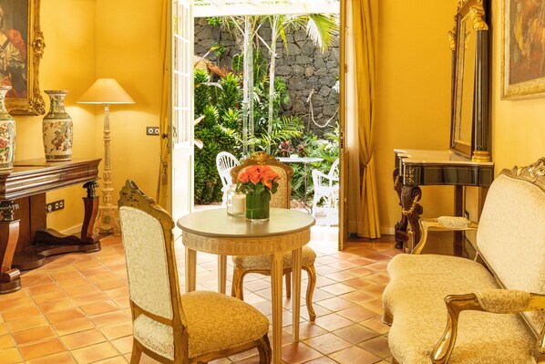 Una habitación con una mesa de comedor, dos sillas, un sofá y una mesita con un jarrón. Hay una puerta que da a un espacio al aire libre con plantas y un muro de piedra.